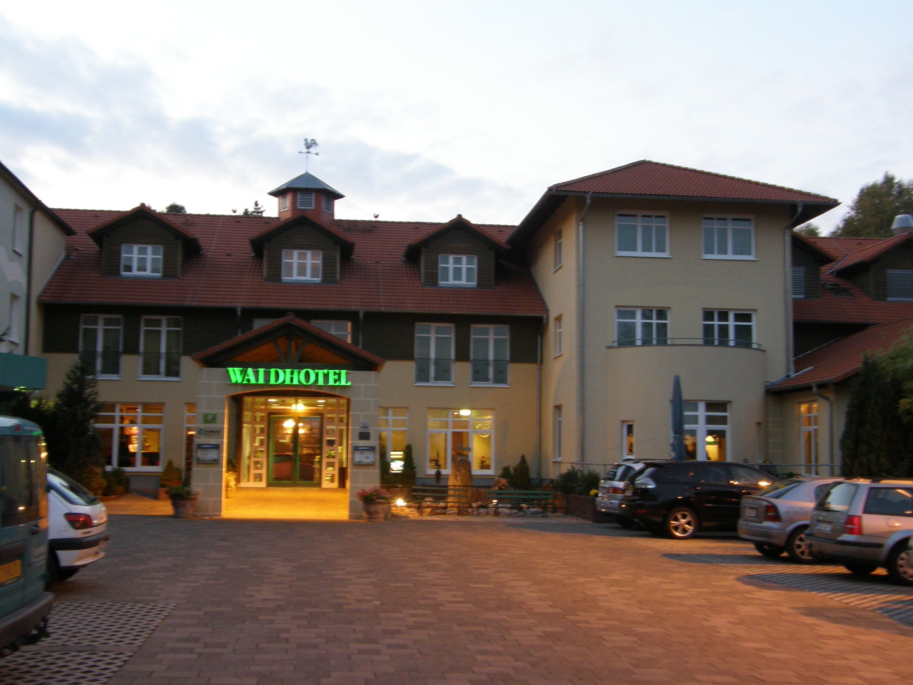 Un hotel de estilo arquitectónico europeo tradicional, con una entrada destacada que incluye un letrero, varias ventanas y un estacionamiento con coches.