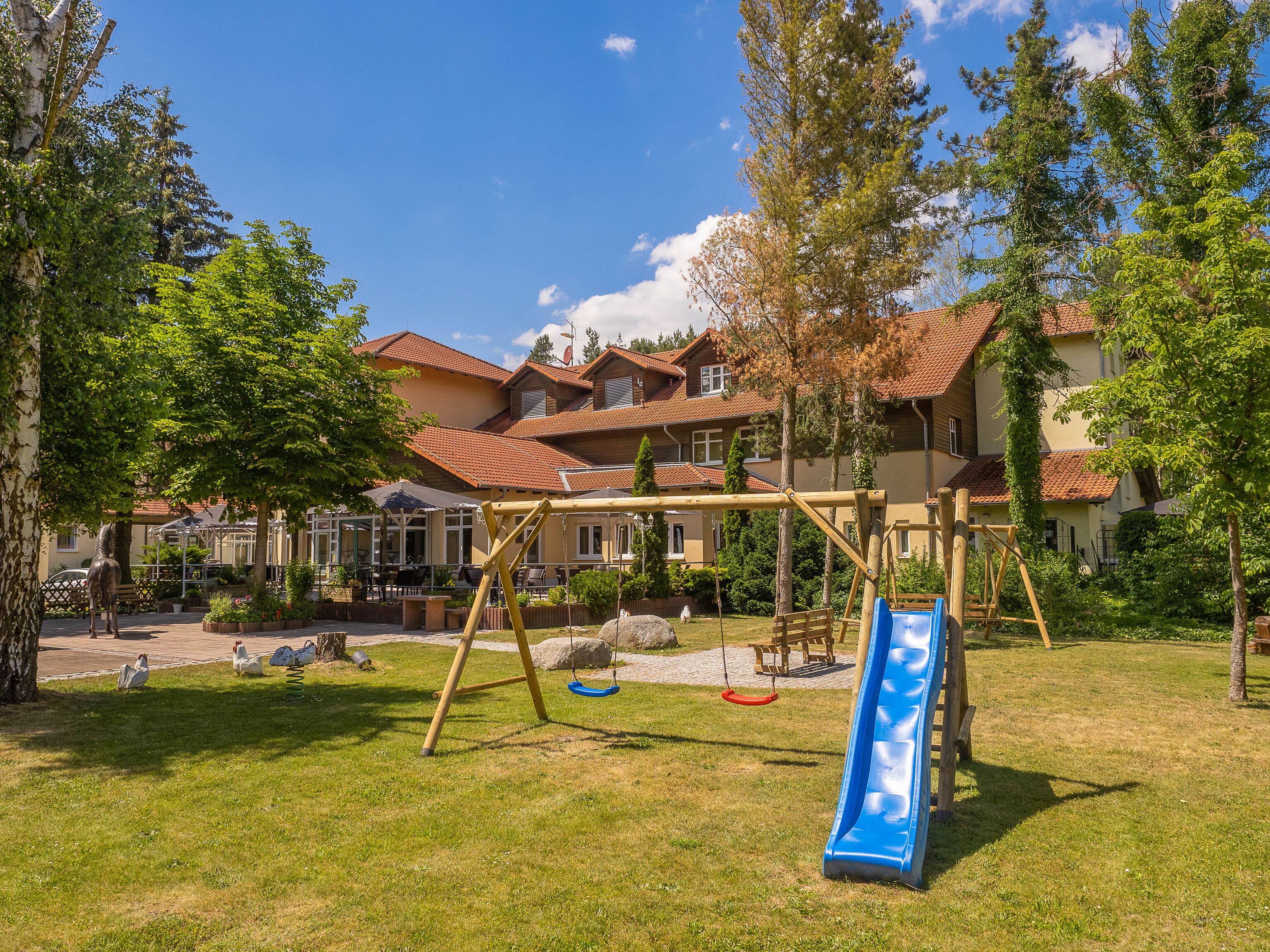 A playground with a slide and swings, surrounded by trees and a building with a red-tiled roof.