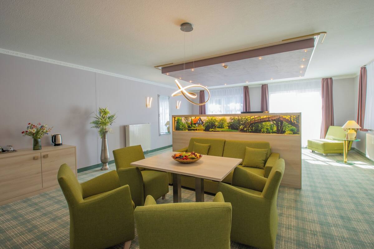 A modern hotel room with a dining area, green upholstered chairs, a table with a fruit bowl, a large window with curtains, and a unique ceiling light fixture.