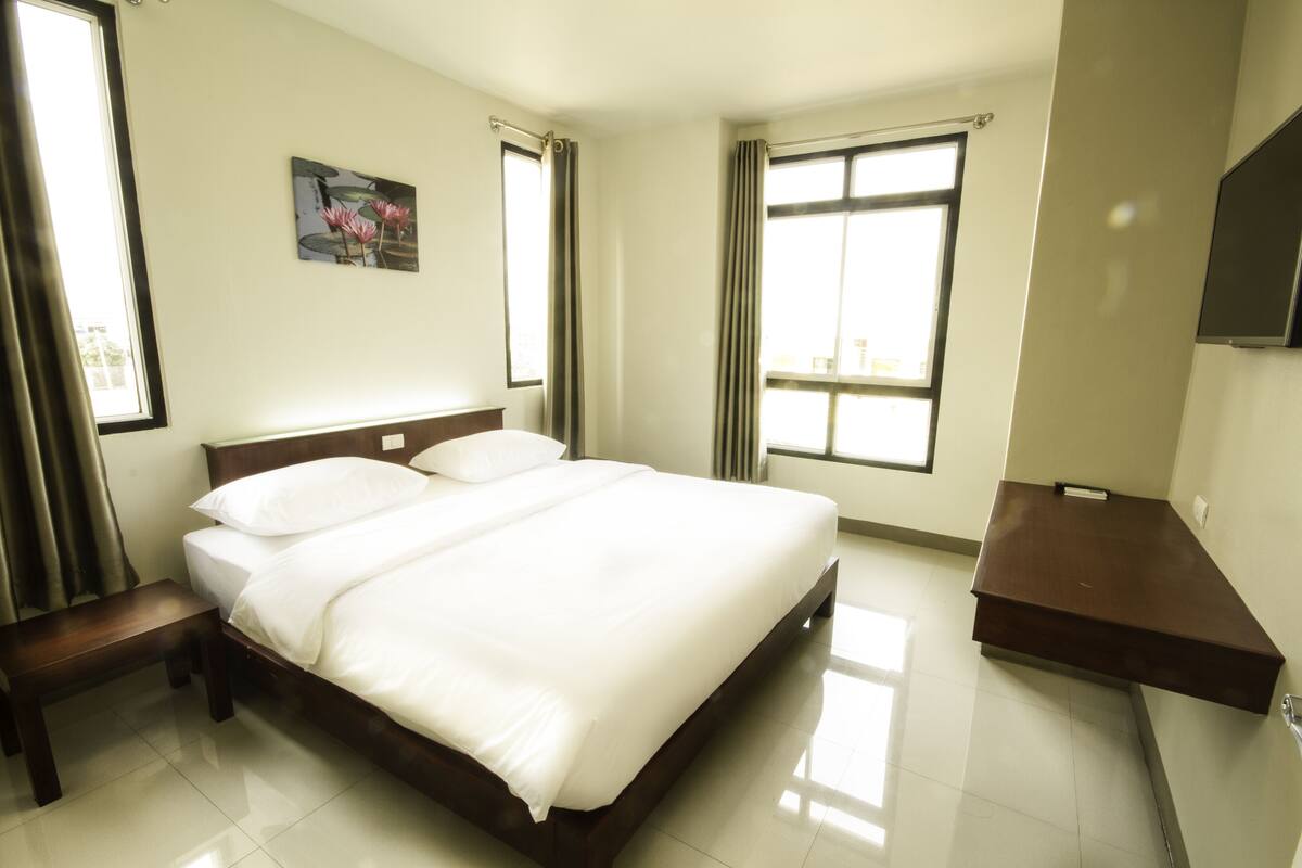 Een hotelkamer met een groot bed, een nachtkastje, een bankje en een aan de muur bevestigde televisie.