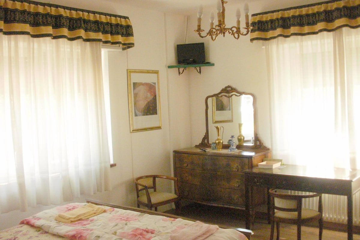 A bedroom with a bed, dresser, chairs, a mirror, and a chandelier.