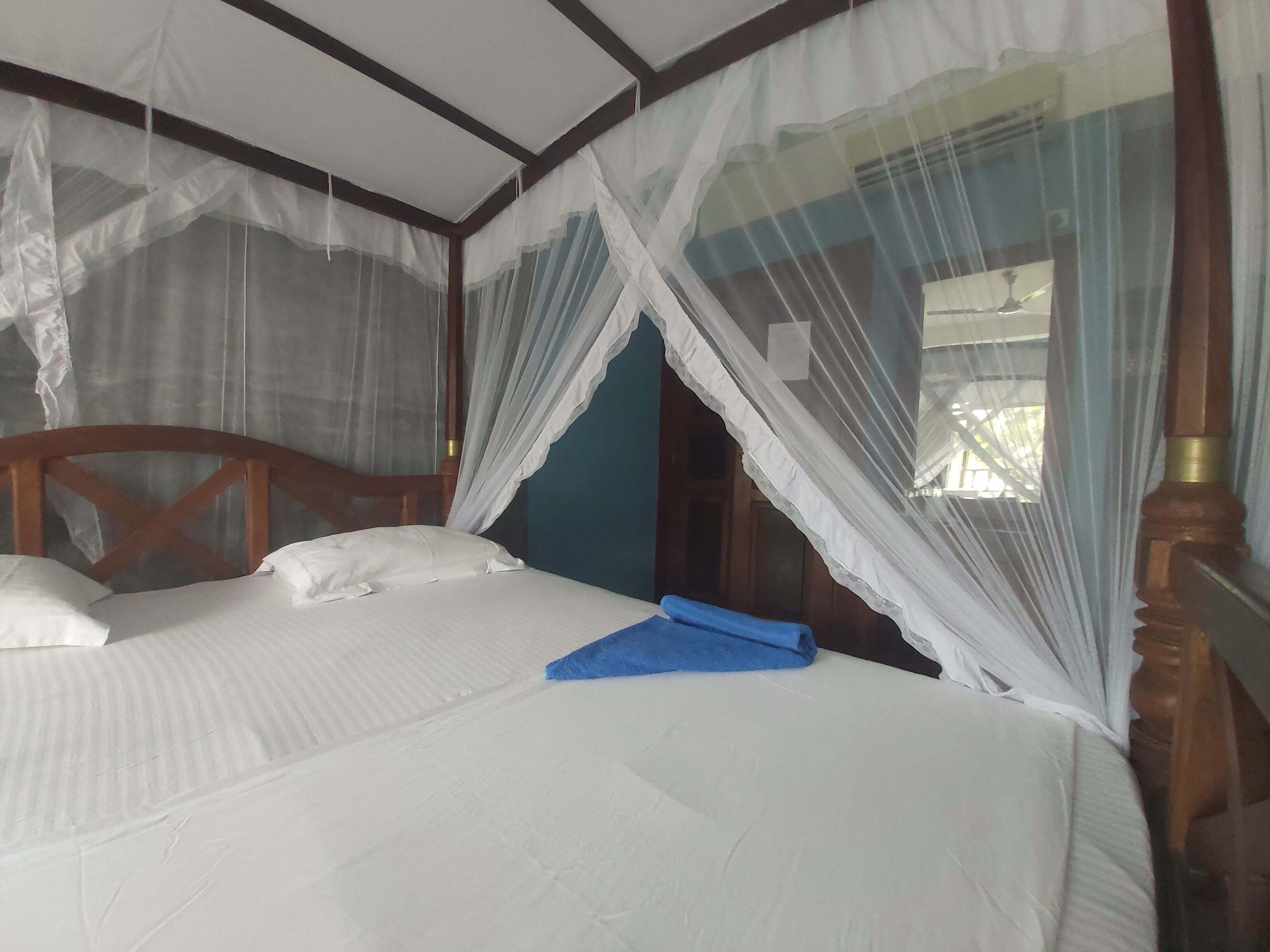 A bed with white linens and a blue towel, a wooden headboard, and a mosquito net.