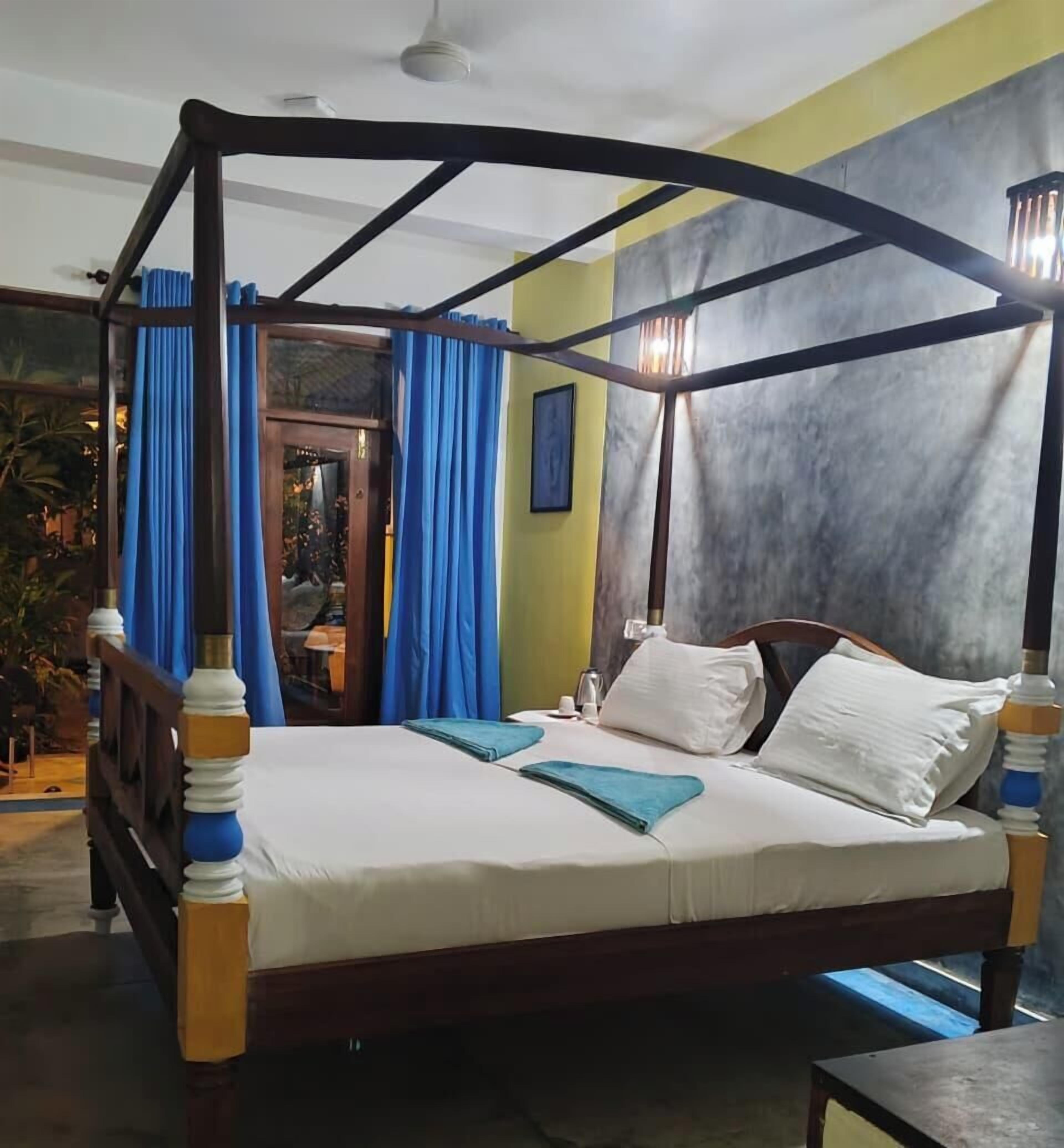 A four-poster bed with a wooden frame and white bedding, a small bedside table, and a view of the outdoors through a glass door.