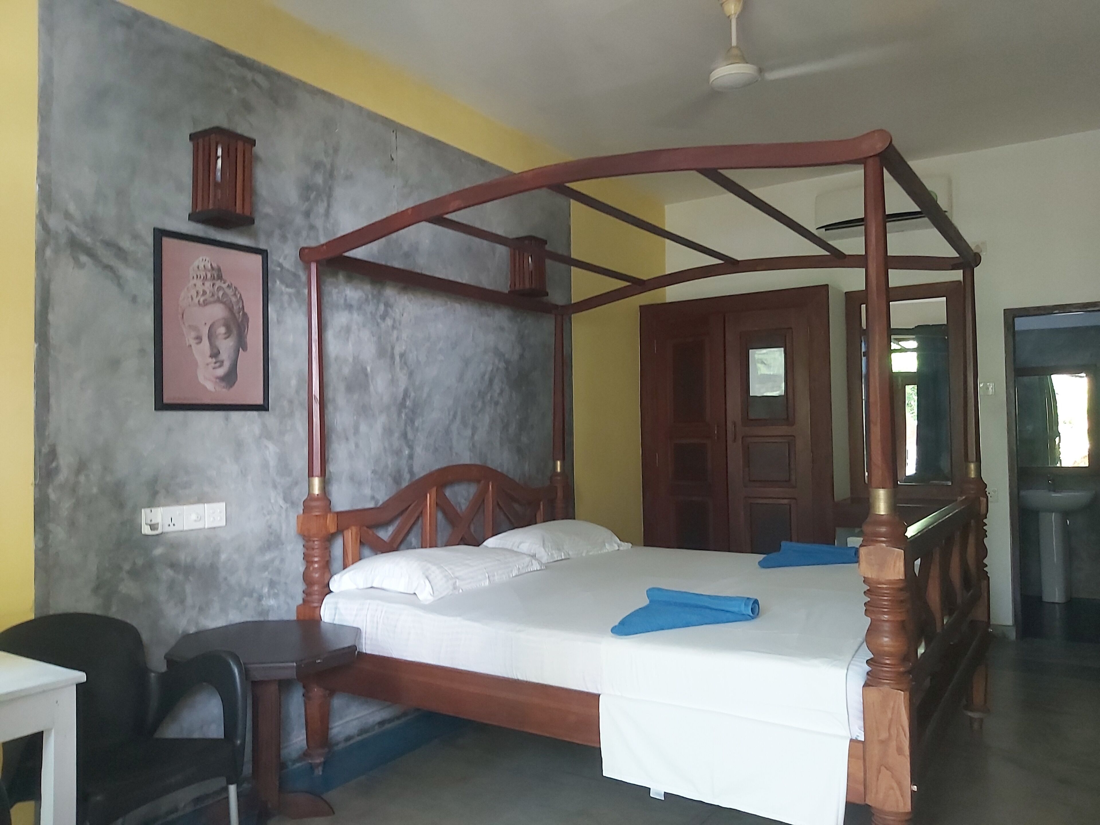 A four-poster bed with a wooden frame, a white mattress, and blue towels. There is a desk with a chair, a wall-mounted shelf with a statue, and a door with a mirror.