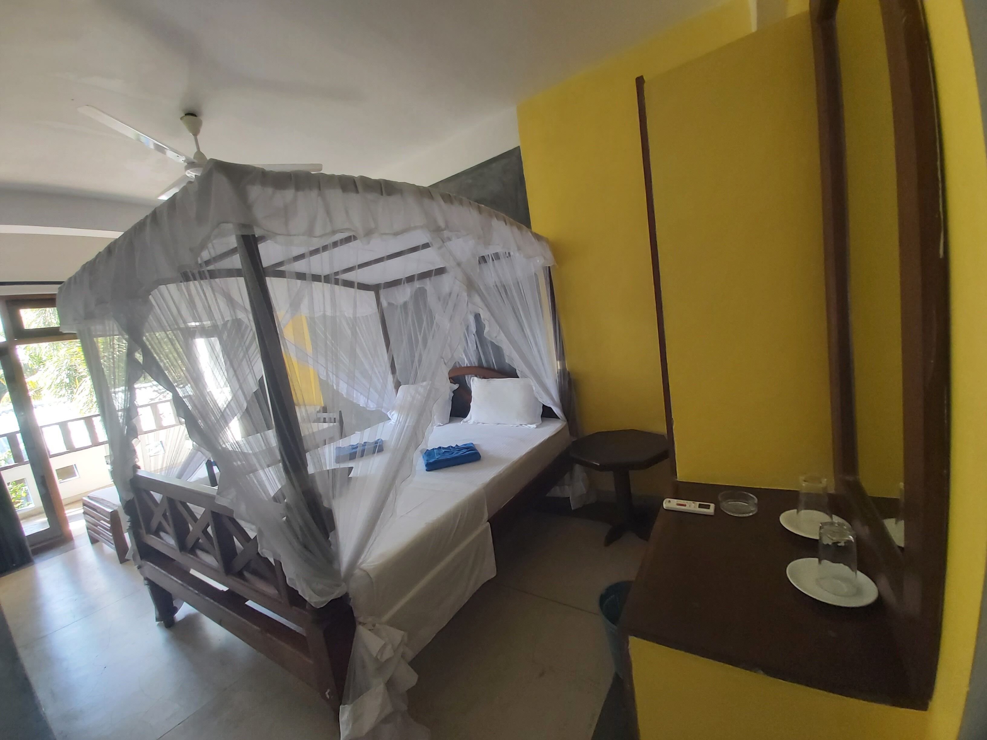 A room with a bed covered by a mosquito net, a bedside table with a thermometer, and a chair.