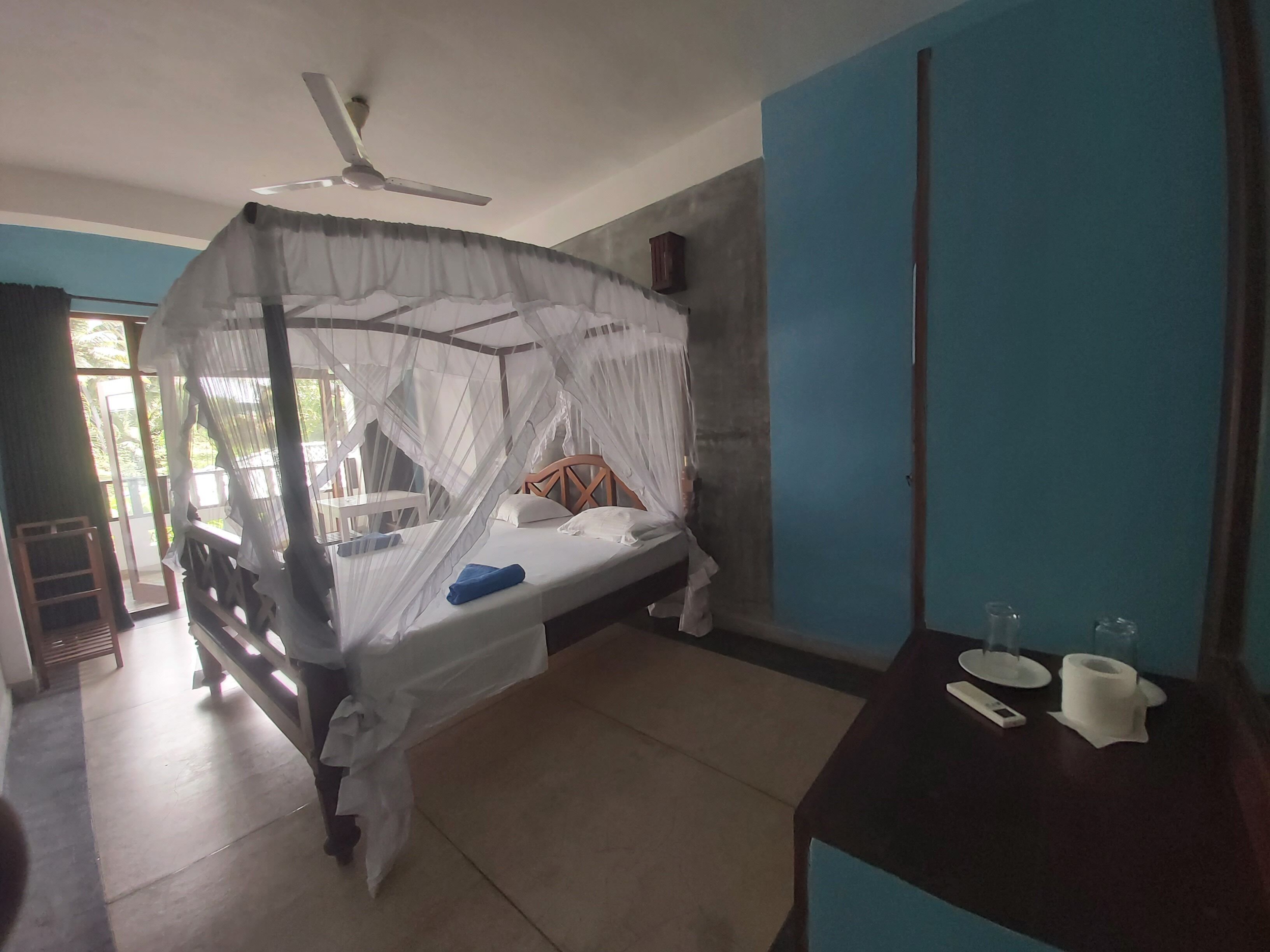 A bedroom with a bed under a mosquito net, a bedside table with a remote control and a glass, and a wardrobe.