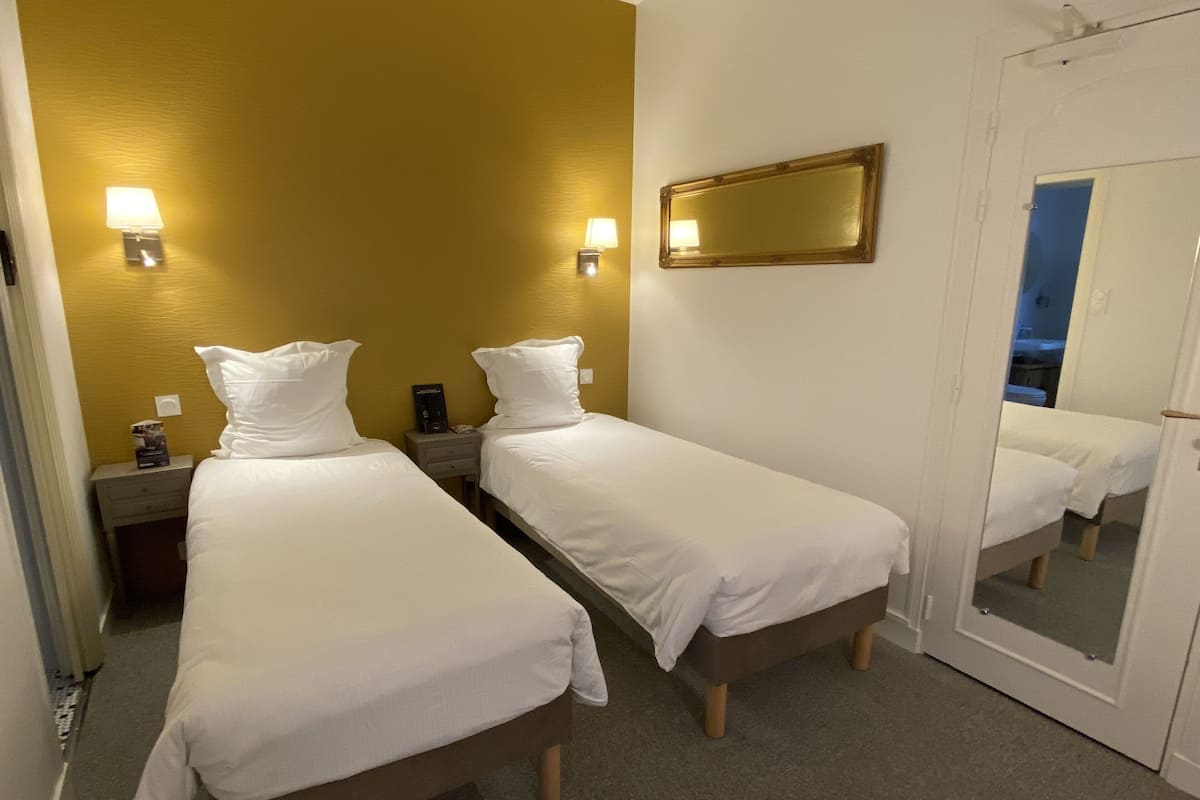 A hotel room with two single beds, a bedside table with a lamp, a mirror, and a door leading to another room.