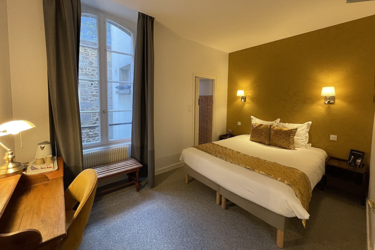 A hotel room with a bed, bedside table, lamp, and a view of a stone building through a window.