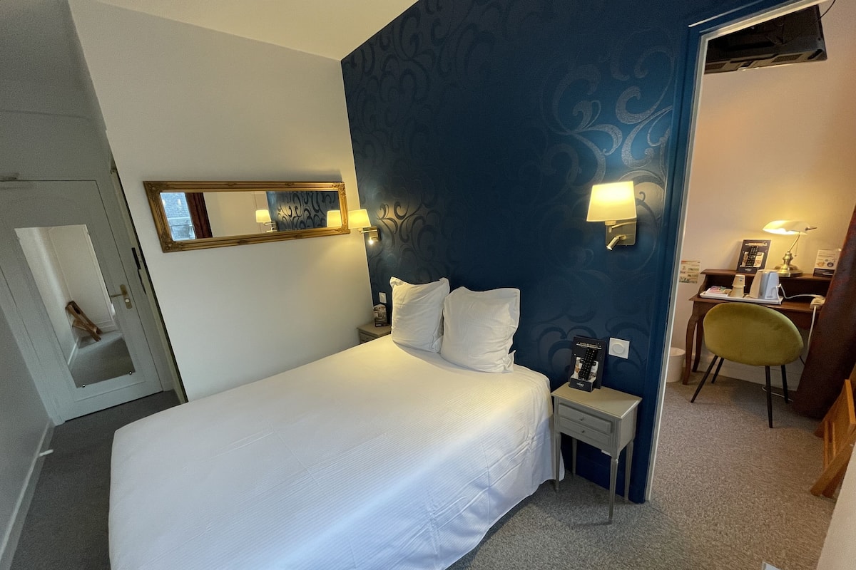 A hotel room with a large bed, a desk with a chair, a small table with a lamp, and a wall-mounted mirror.
