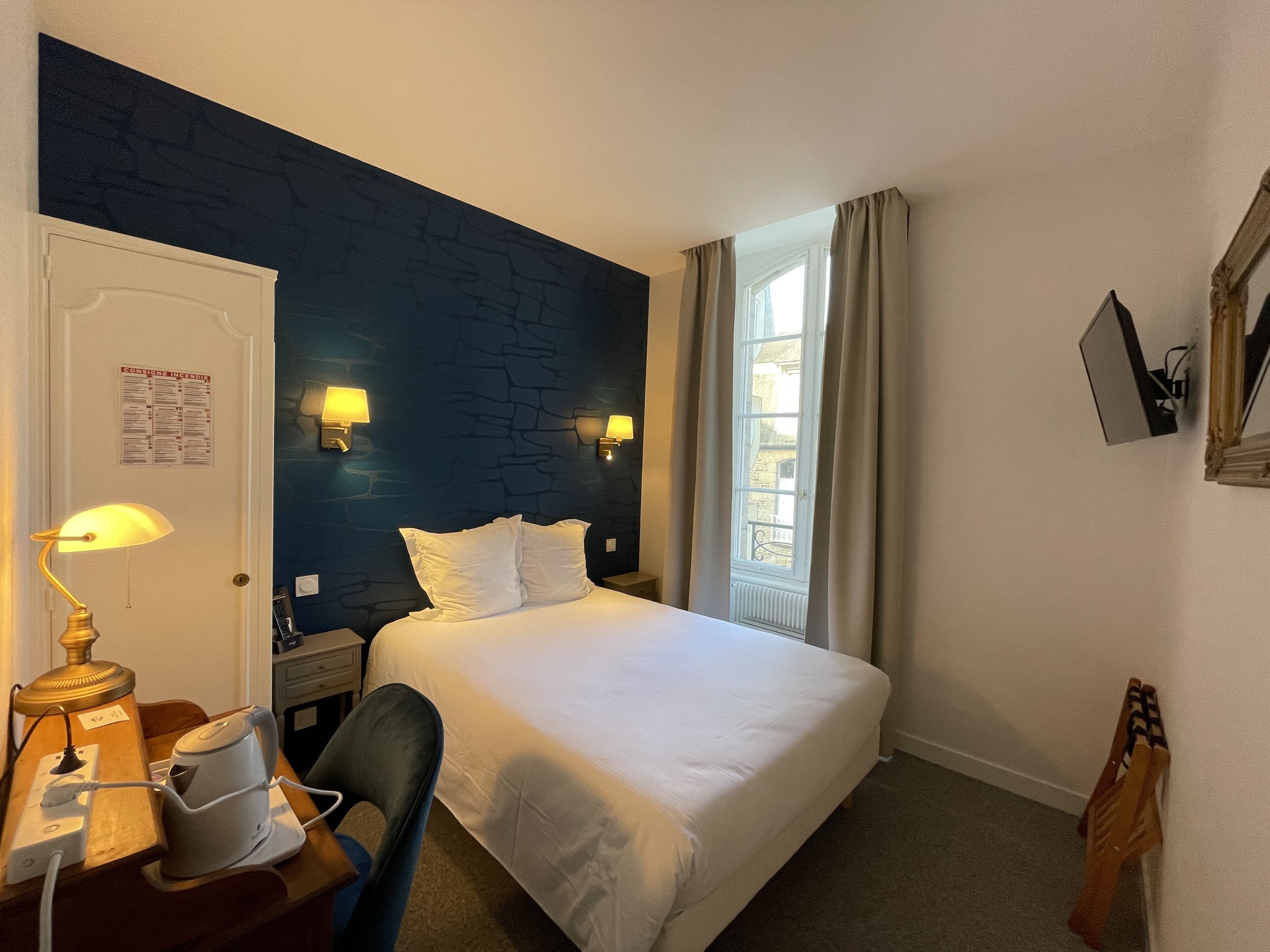 Ein Hotelzimmer mit Bett, Schreibtisch mit Lampe, Sessel, Nachttisch und einem Fenster mit Vorhängen.