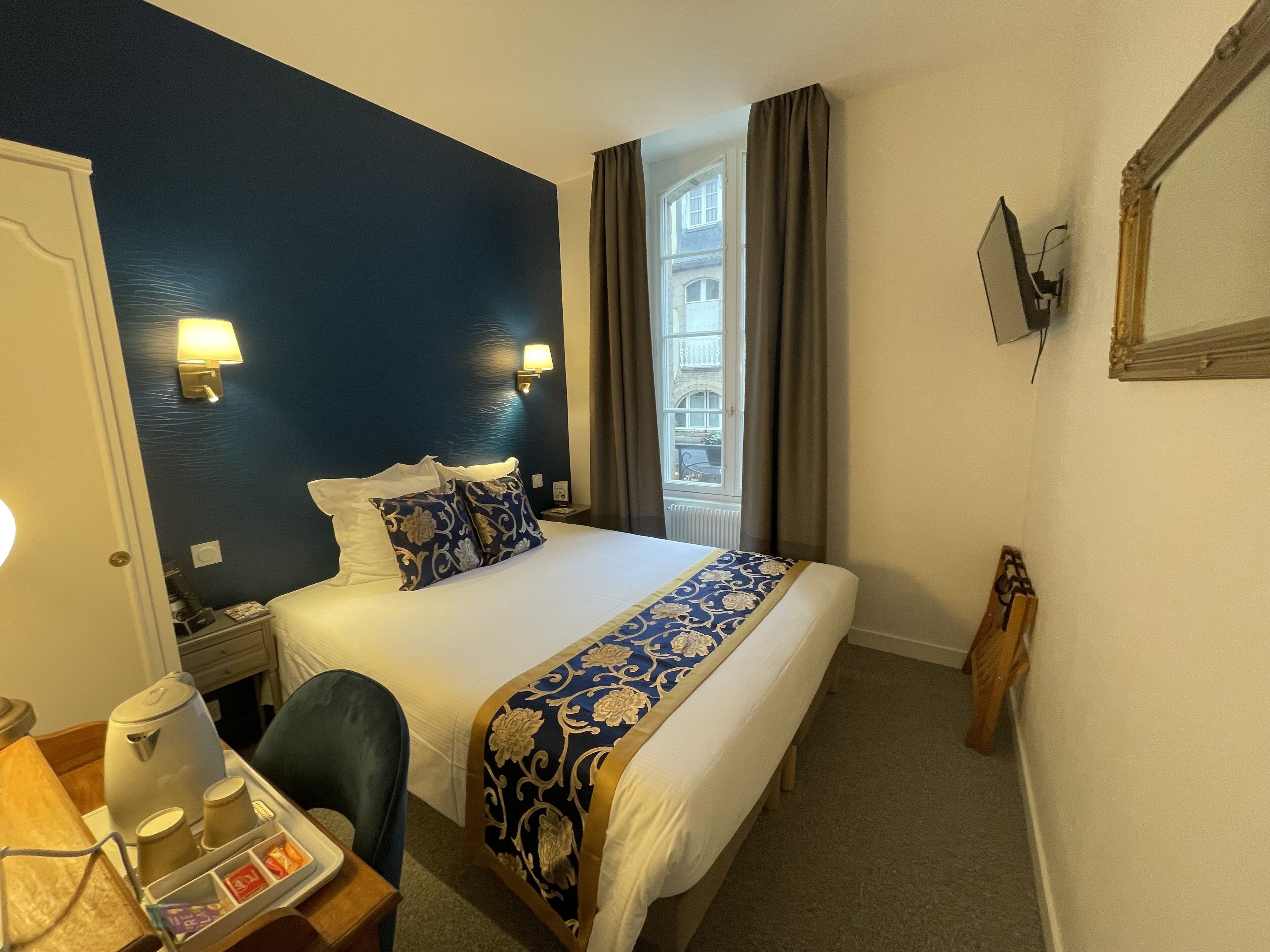 A hotel room with a large bed, a desk with a kettle and cups, a chair, and a view of a building through the window.