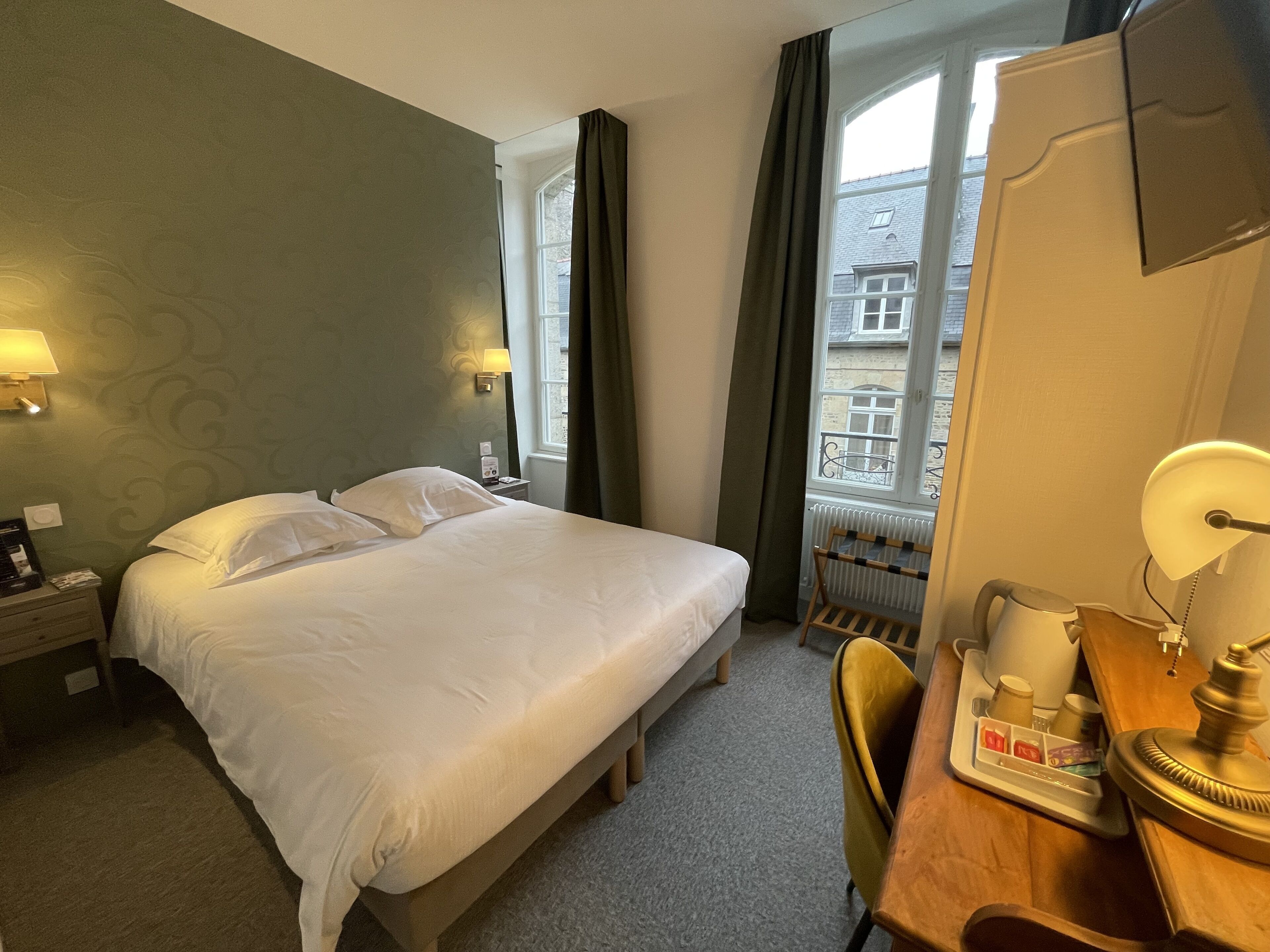 Ein Hotelzimmer mit einem Bett, einem Schreibtisch mit Lampe, einem Sessel und einem Fenster mit Vorhängen.