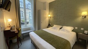 A hotel room with a bed, a desk, a chair, a window with curtains, a stone wall, and two bedside lamps. - Balzac Hôtel (Fougeres)