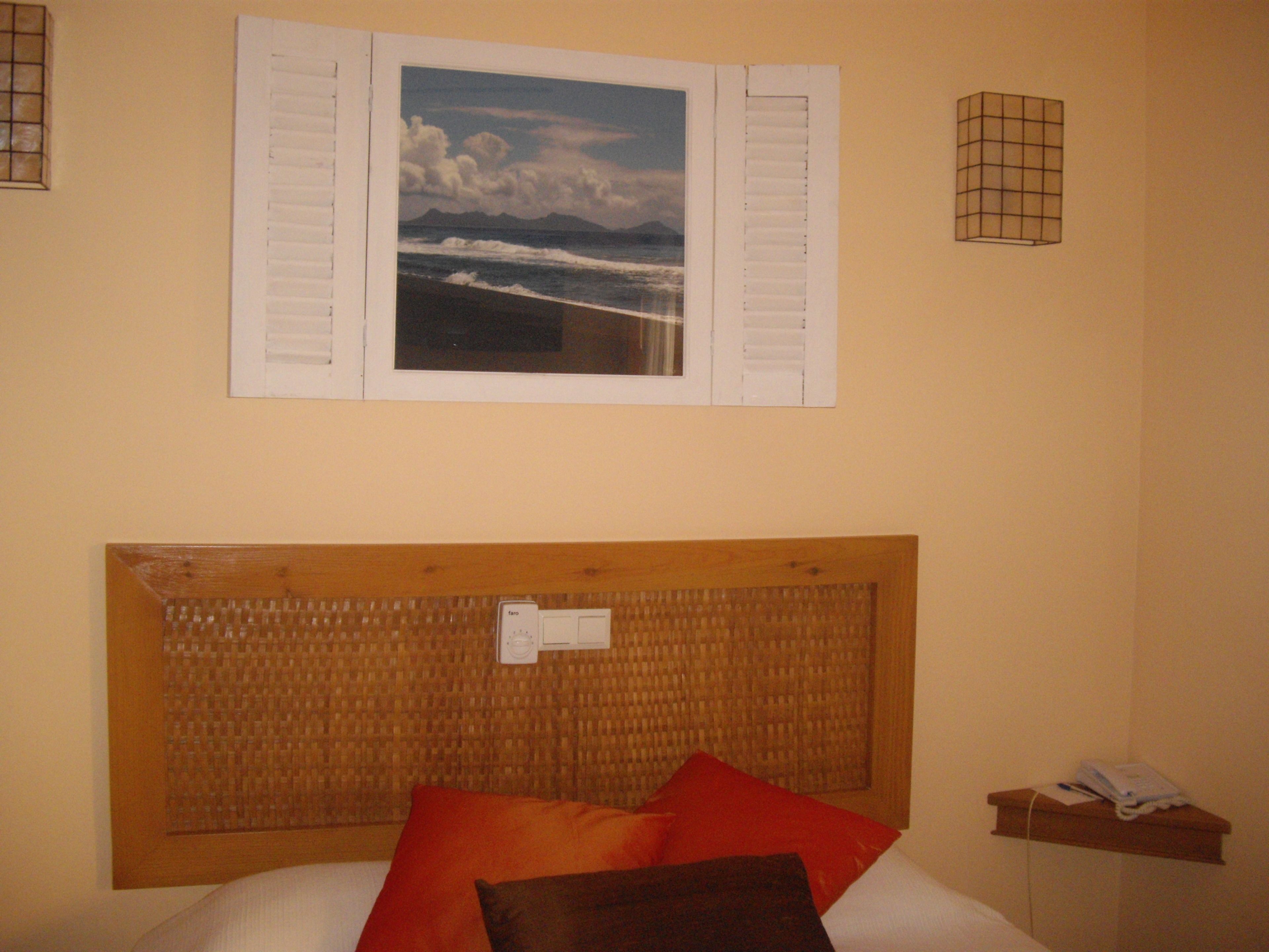 A bedroom with a bed, a headboard, a wall-mounted picture of a beach, and a bedside table with a lamp.