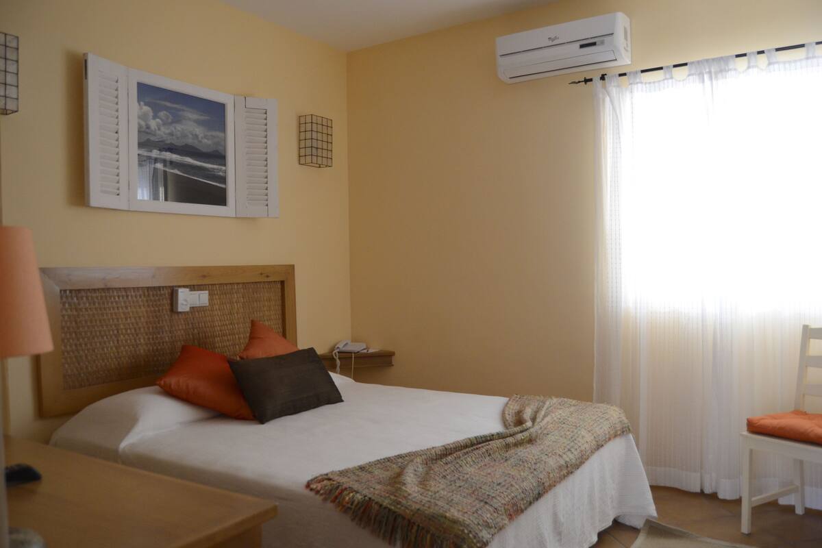 A hotel room with a bed, a bedside table, a lamp, an air conditioner, and a chair.