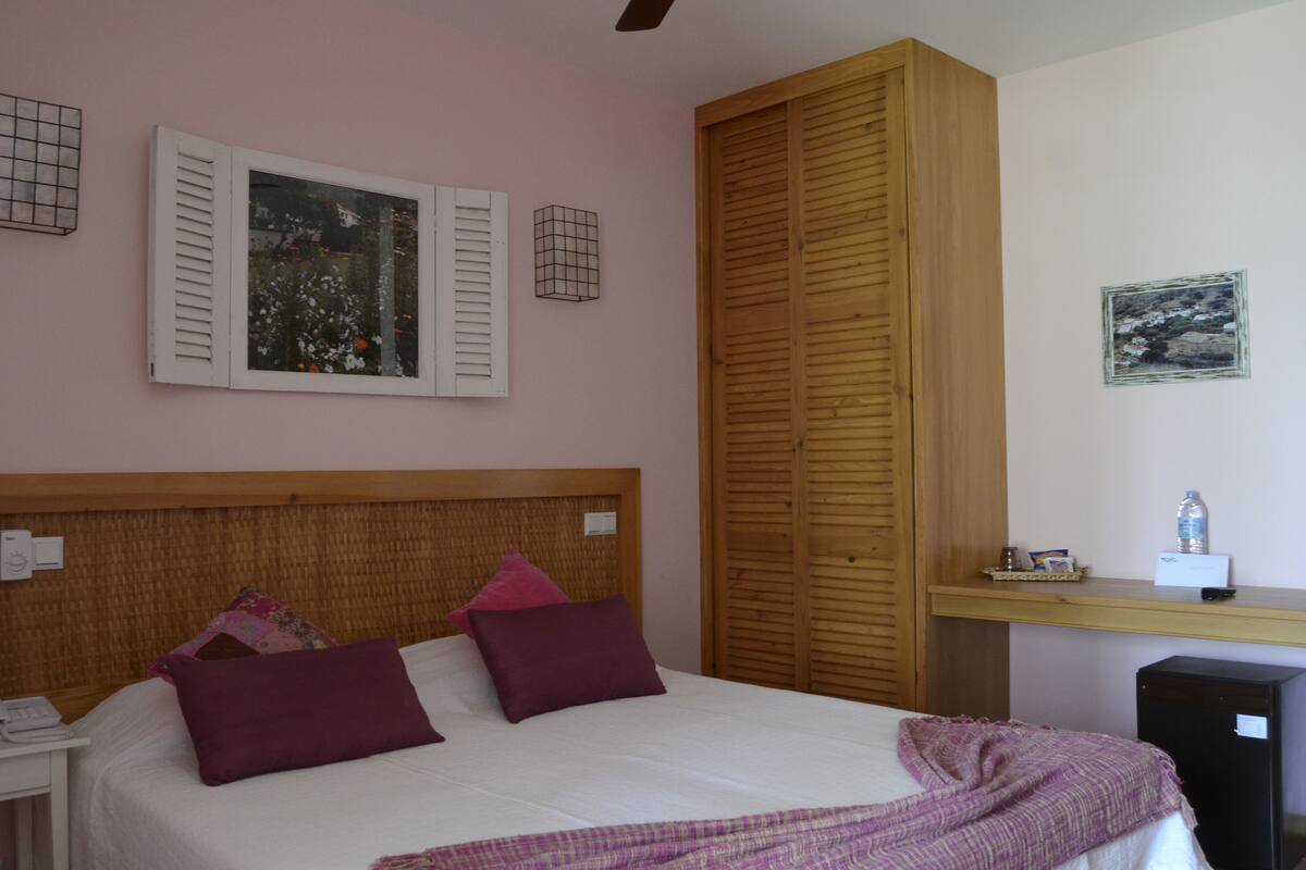 A bedroom with a bed, a wooden wardrobe, a nightstand, a desk, and a small refrigerator.