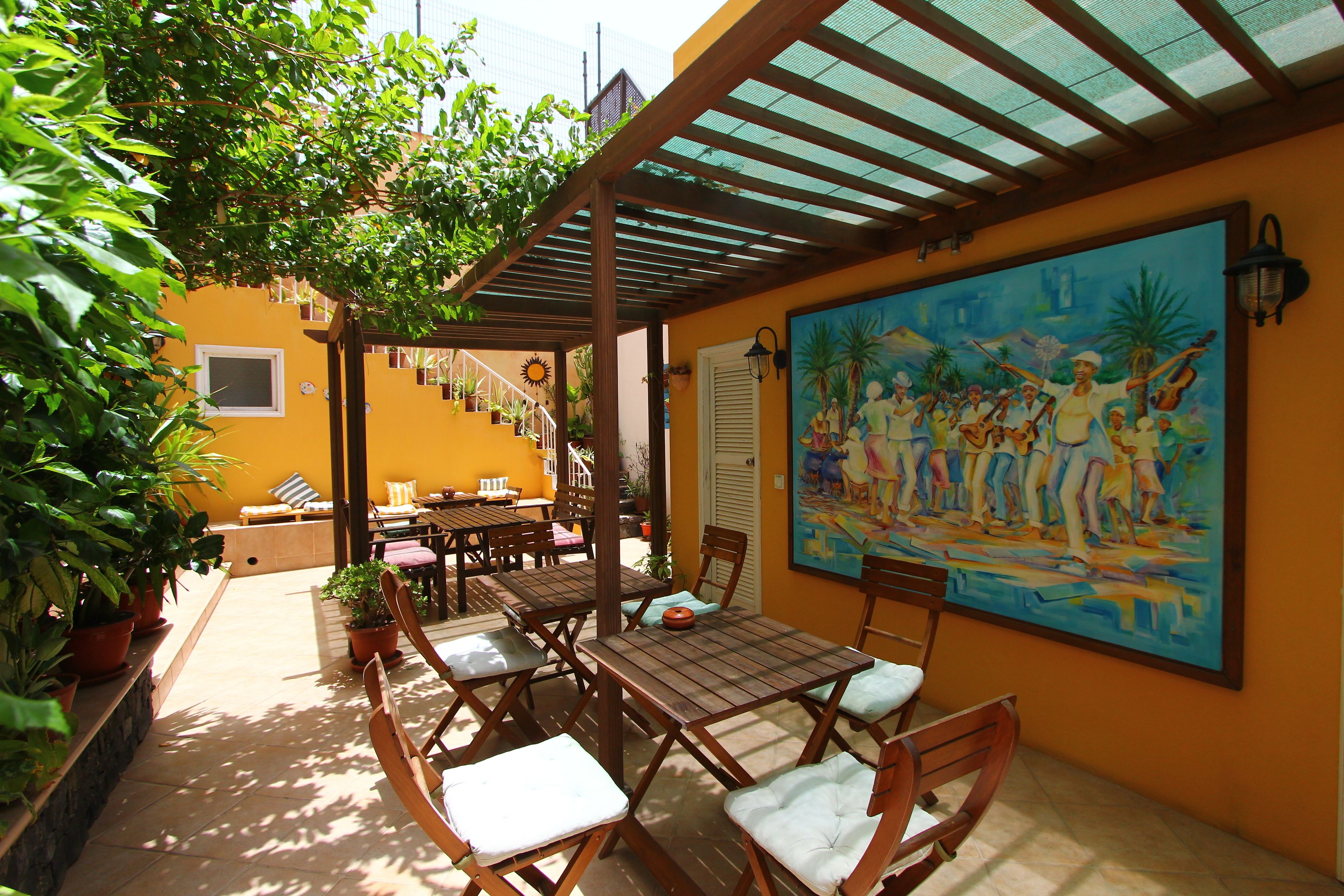 An outdoor patio with wooden tables and chairs, potted plants, and a mural on the wall.