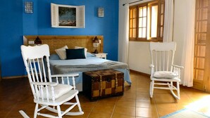 Un dormitorio con cama, cabecera de madera, dos mecedoras, un aparador y una ventana con cortinas.