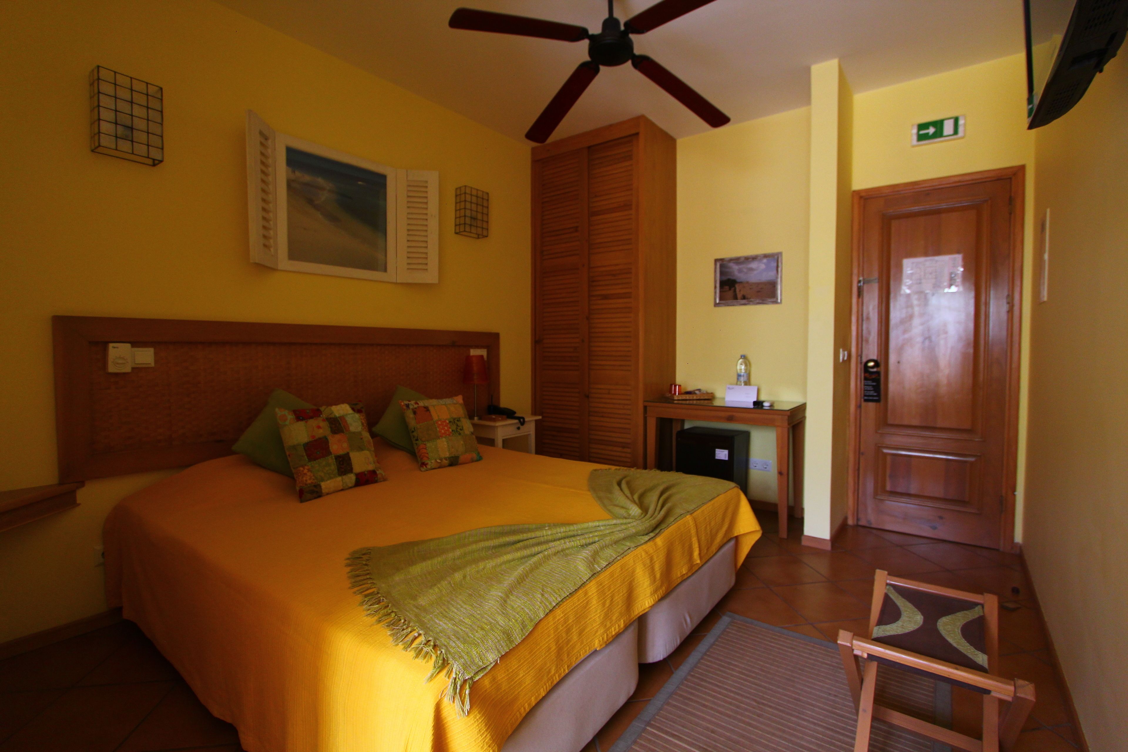 Habitación de hotel con cama, escritorio, silla, ventilador de techo y puerta con letrero de salida.
