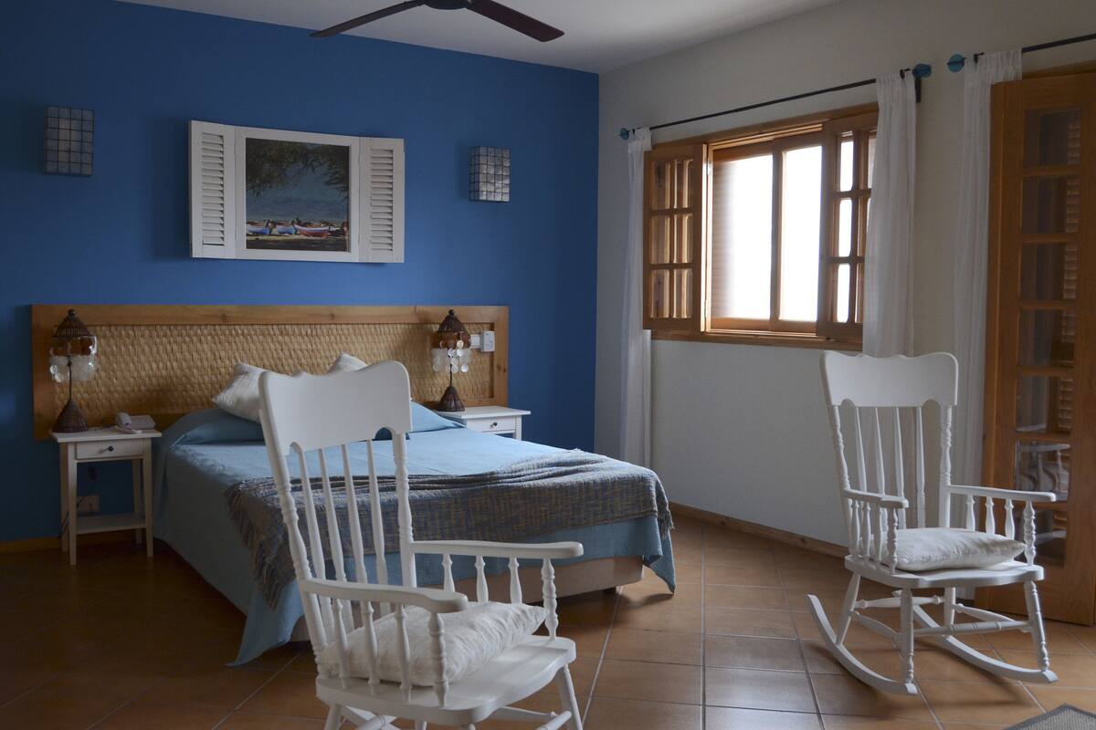 A bedroom with a bed, two nightstands, a rocking chair, a wooden headboard, and a window with curtains.