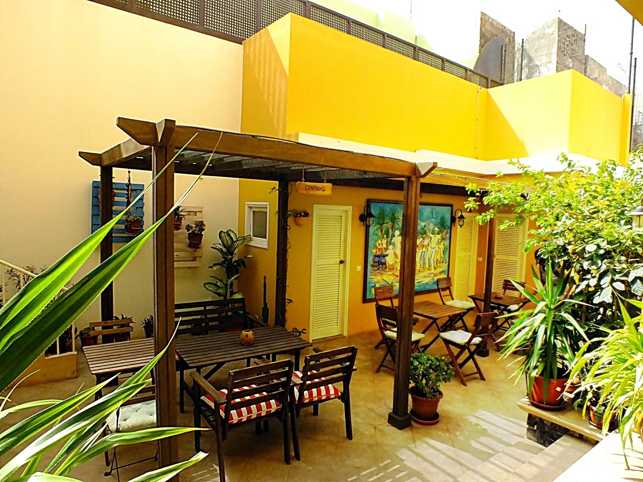 An outdoor patio with wooden tables and chairs, potted plants, and a yellow wall with a door labeled "CANTINA".