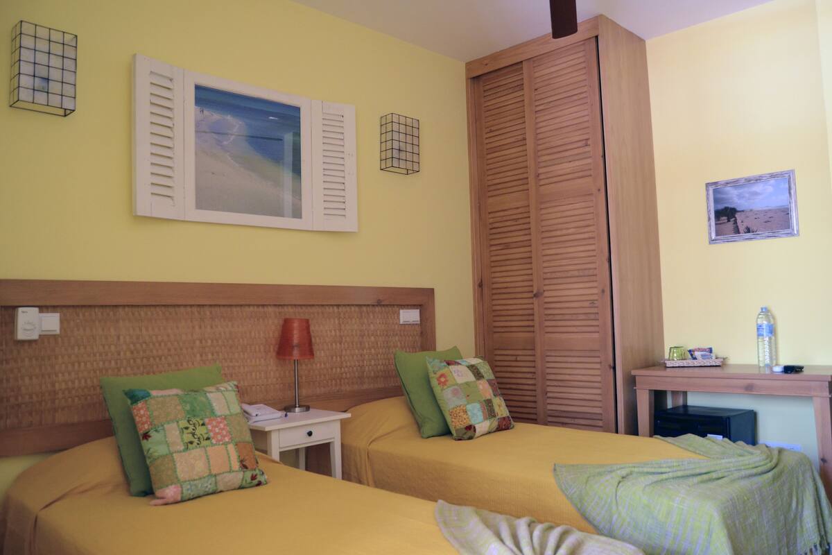 A hotel room with two beds, a nightstand, a lamp, a wooden wardrobe, and a small table with a water bottle.