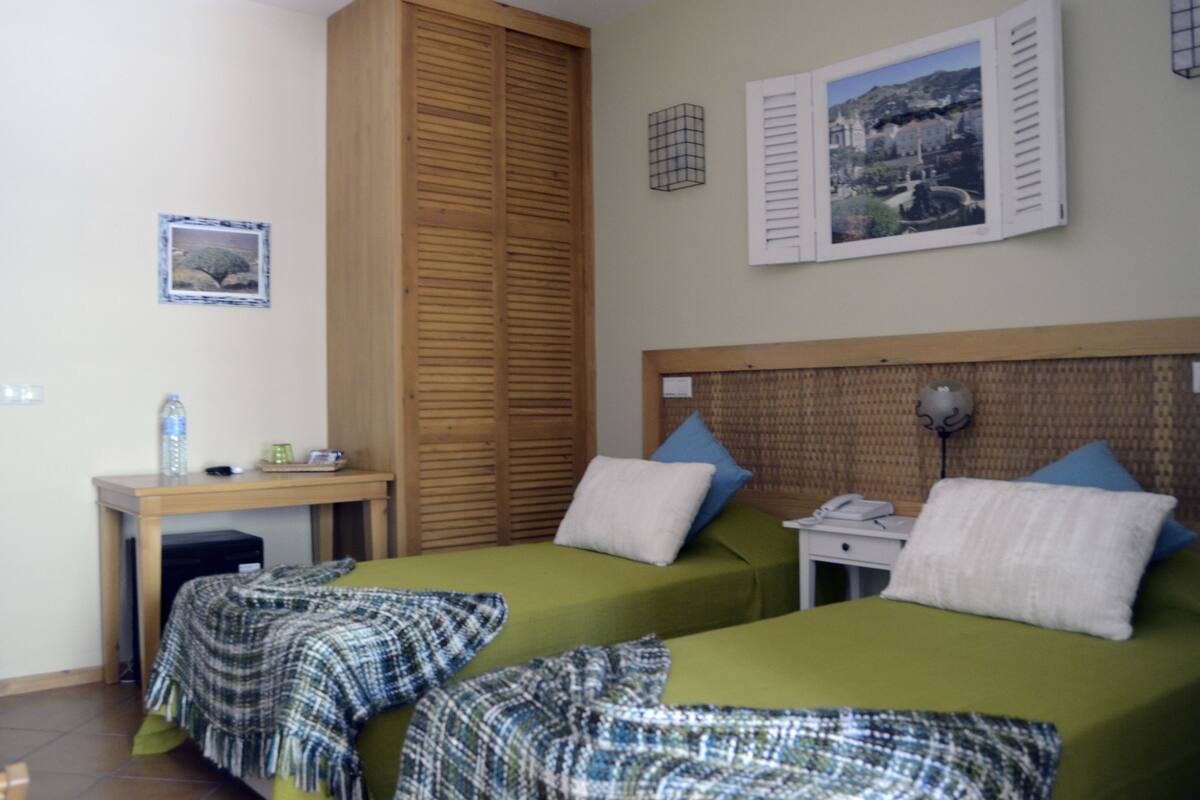 A room with two beds, a wooden wardrobe, a desk with a chair, and a wall-mounted lamp.