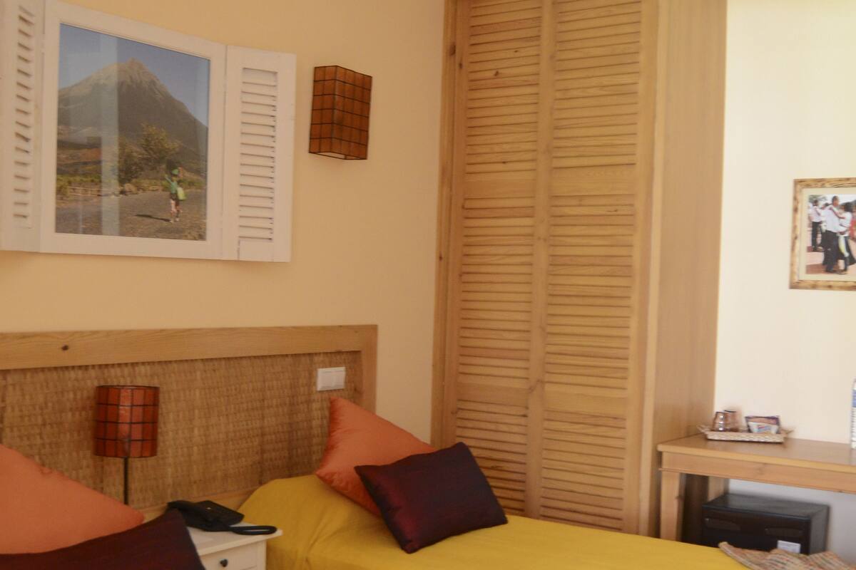 A hotel room with two beds, a ceiling fan, a wooden wardrobe, and a small desk with a lamp.
