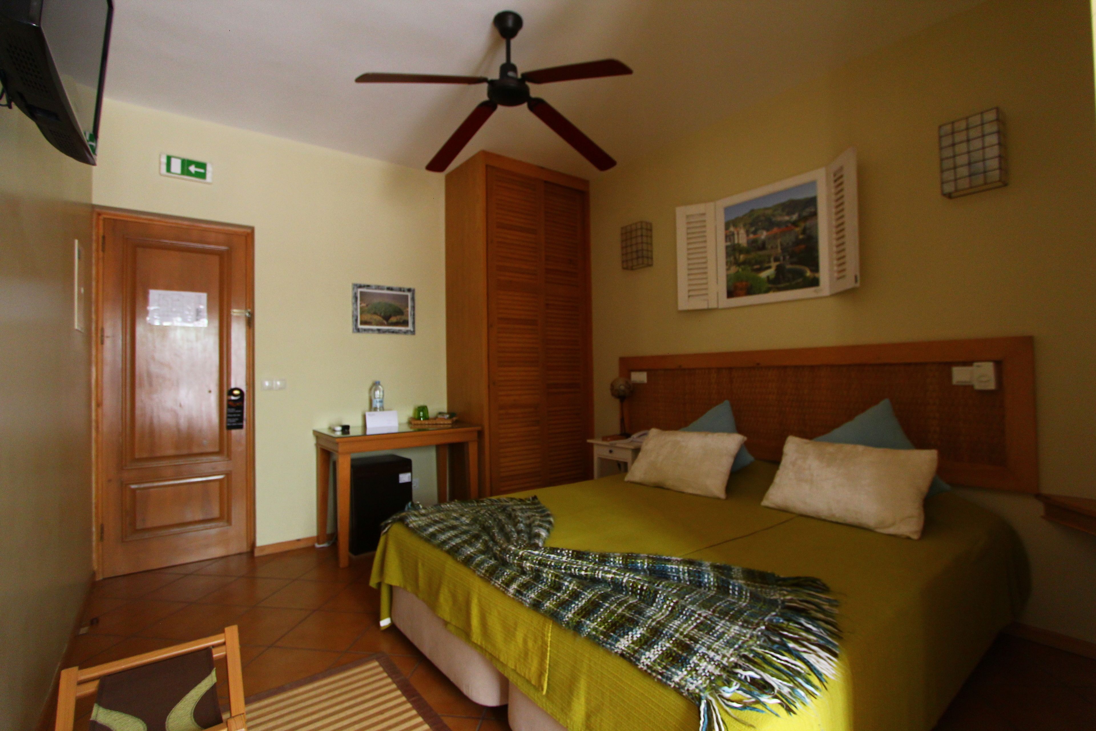 A hotel room with a bed, a desk, a ceiling fan, a door, and a picture on the wall.