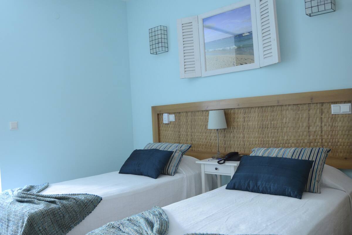 A room with two beds, a wooden headboard, a bedside table with a lamp, and a window with a view of the sea.