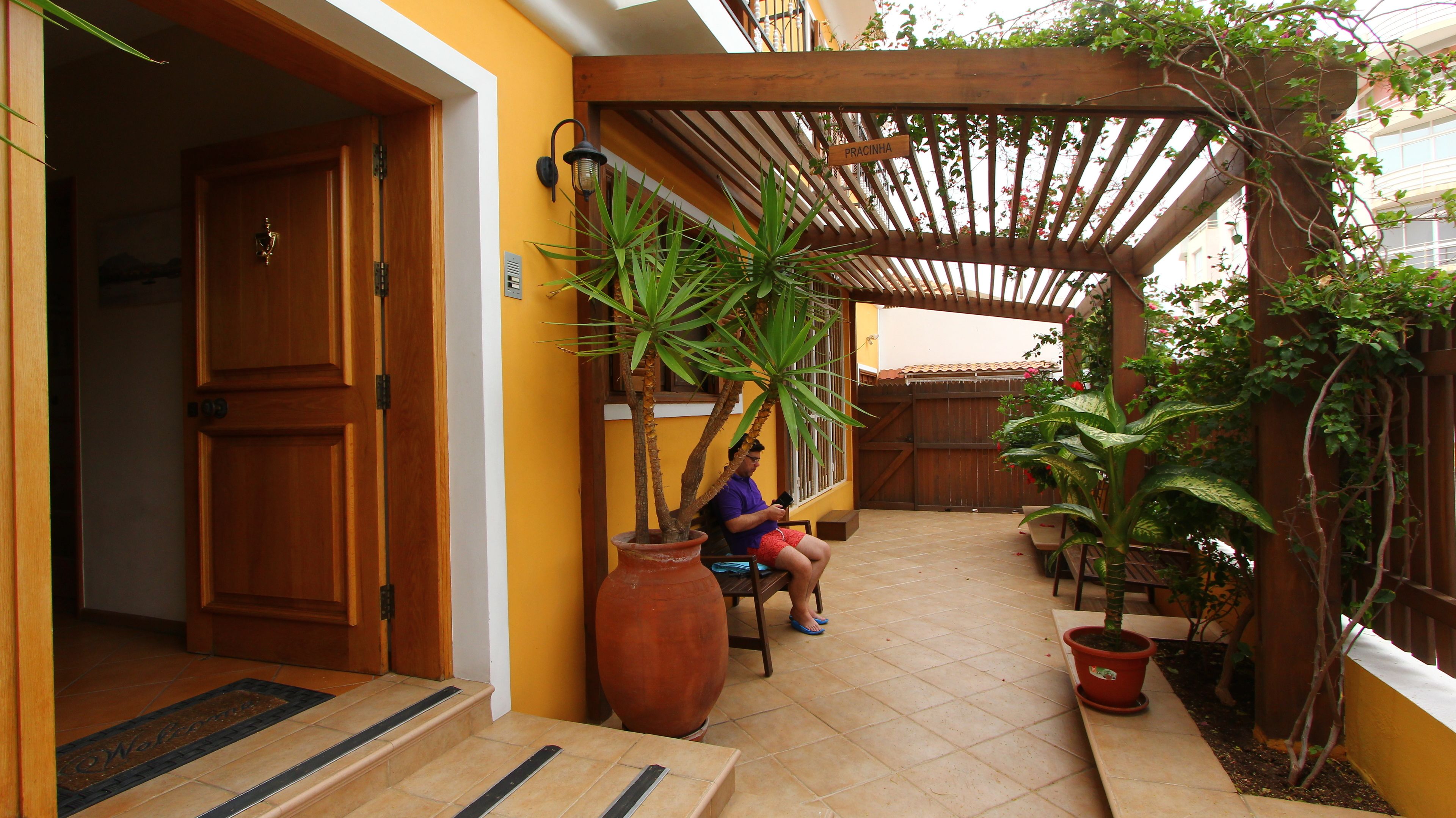 A covered patio area with a wooden pergola, potted plants, and a person sitting on a bench.