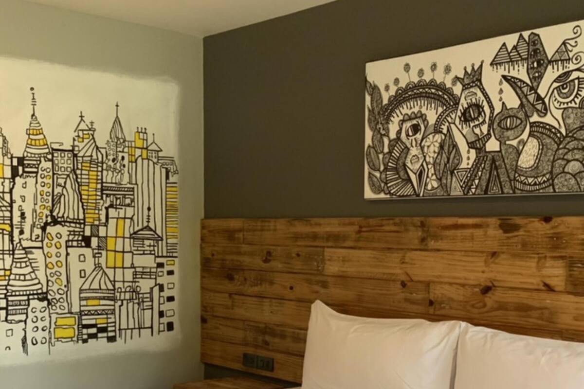 Ein Hotelzimmer mit einem großen Bett, einem hölzernen Kopfteil, einem Stadtbild-Wandgemälde und einem Kunstwerk im Stammesstil an der Wand.