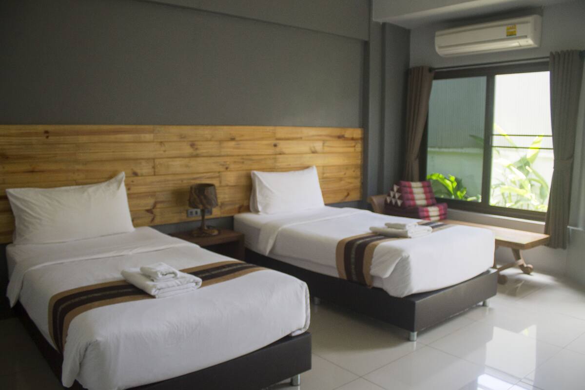 Ein Hotelzimmer mit zwei Betten, einem hölzernen Kopfstück, weißer Bettwäsche, einem kleinen Tisch und einem Fenster mit Vorhängen.