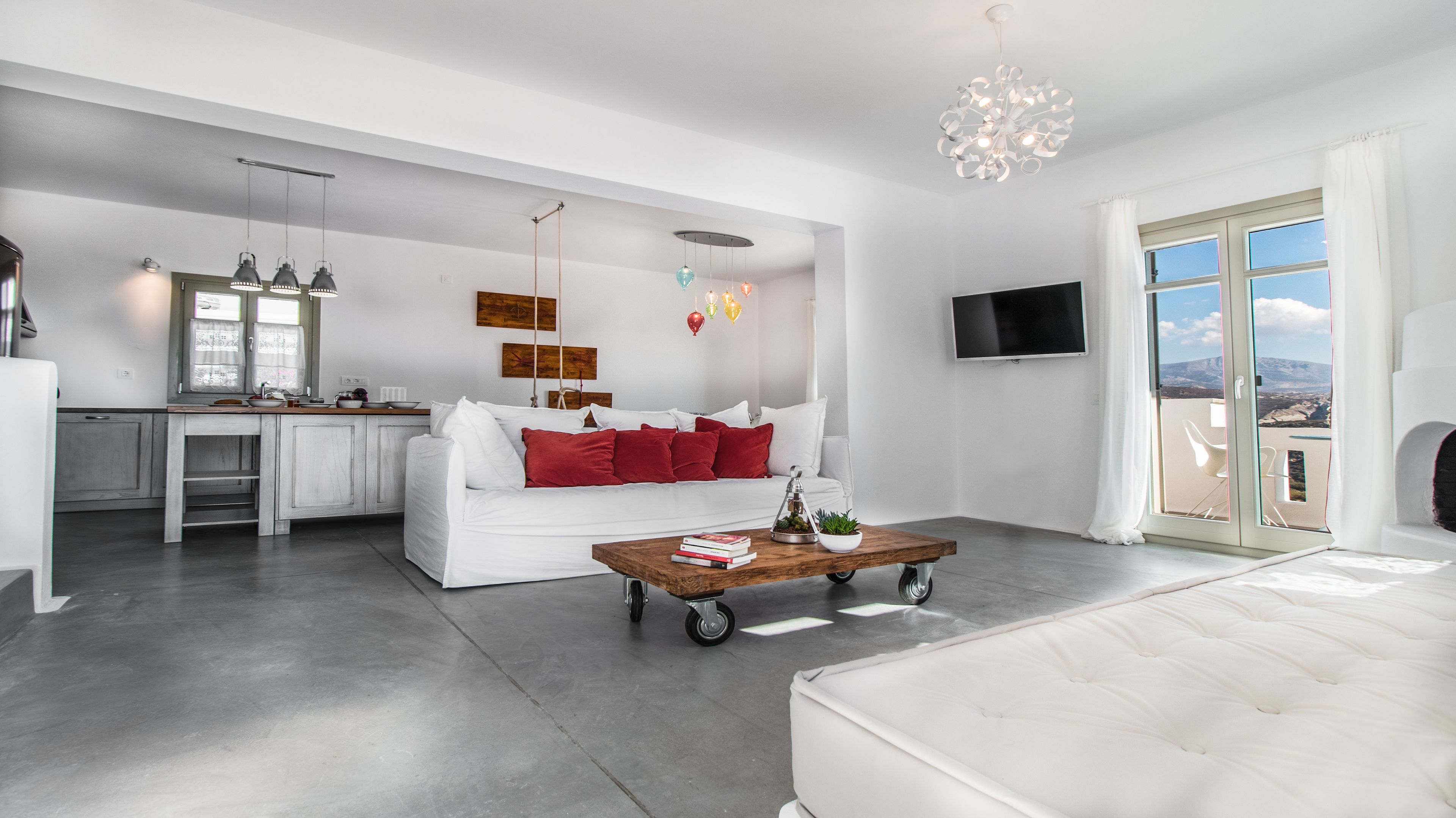 A modern living room with a white sofa, red cushions, a wooden coffee table, and a balcony with a view.