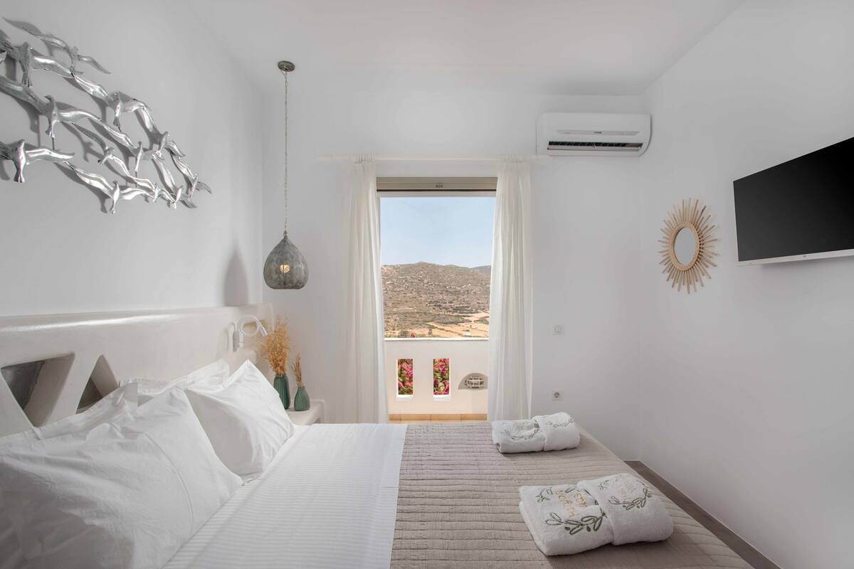 A bedroom with a bed, a view of the landscape, and a TV on the wall.