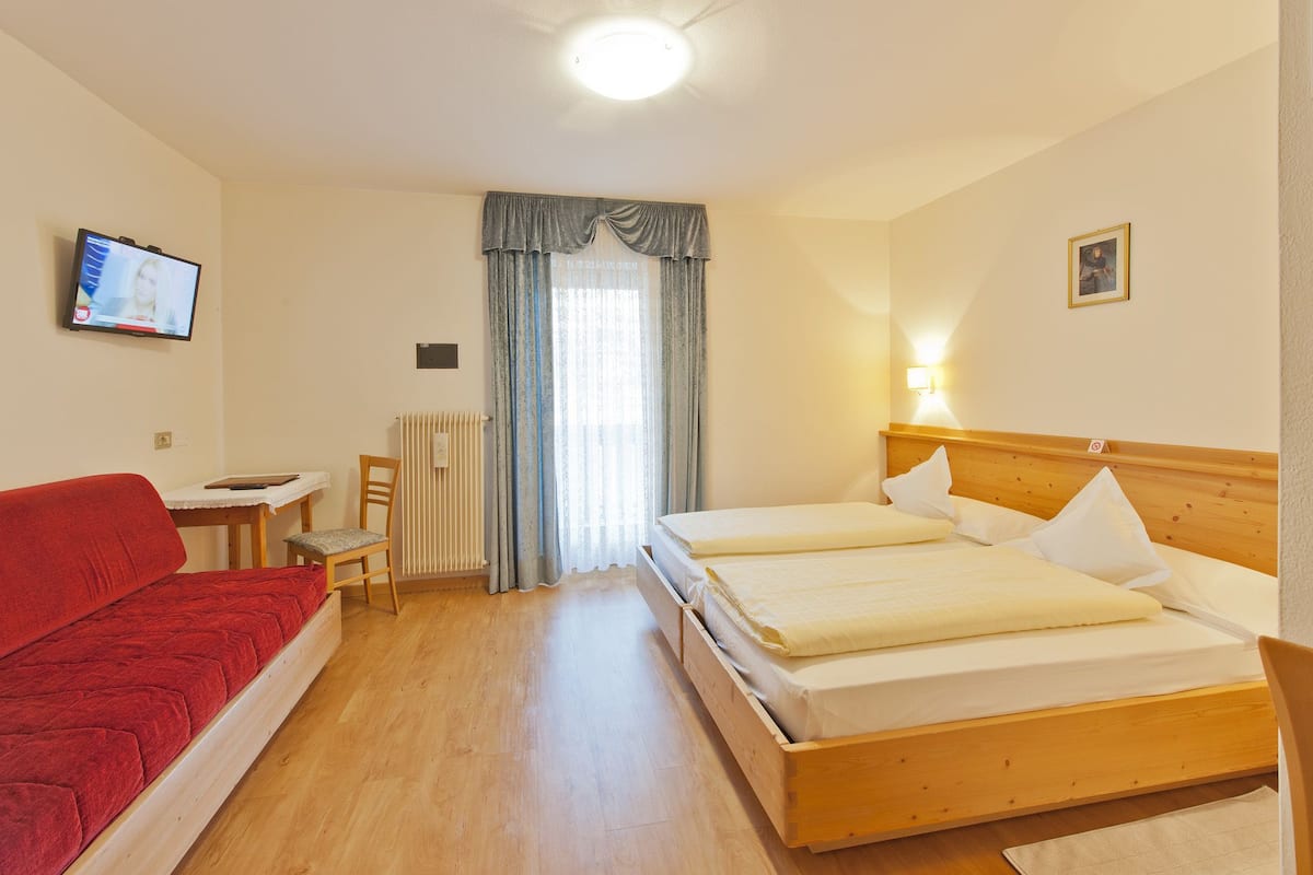 Ein Hotelzimmer mit einem Bett, einem roten Sofa, einem kleinen Tisch, einem Sessel, einem Fernseher und einem Fenster mit Vorhängen.