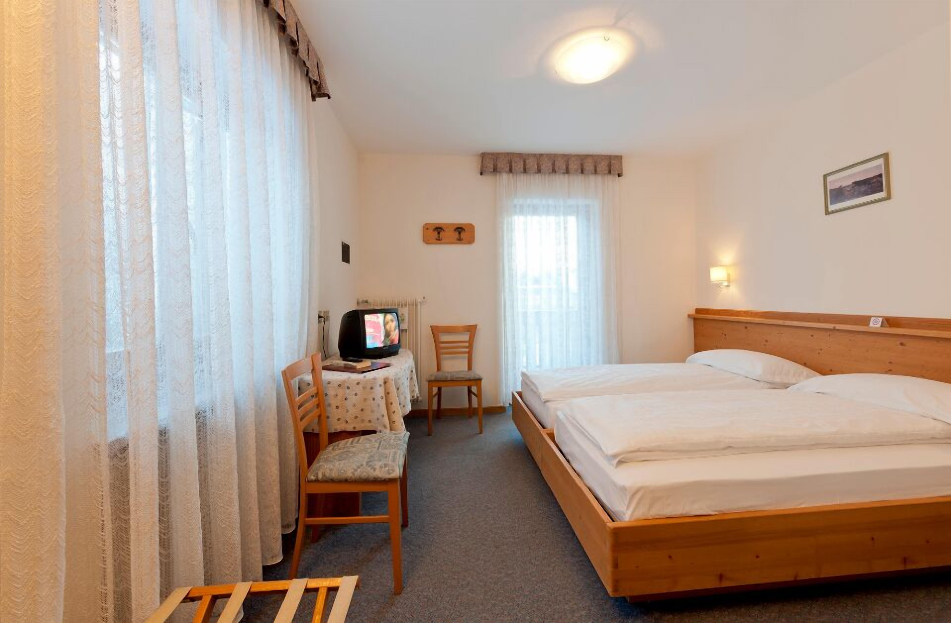 A bedroom with a wooden bed, a desk with a chair, a television, and a window with curtains.