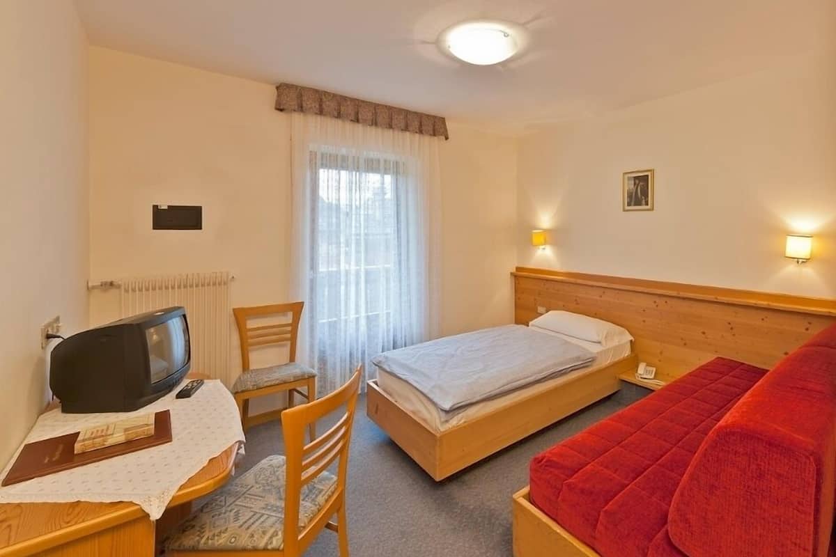 Ein Hotelzimmer mit einem Einzelbett, einem Schreibtisch mit Fernseher, einem Sessel und einem roten Sofa, sowie einem Fenster mit Vorhängen.