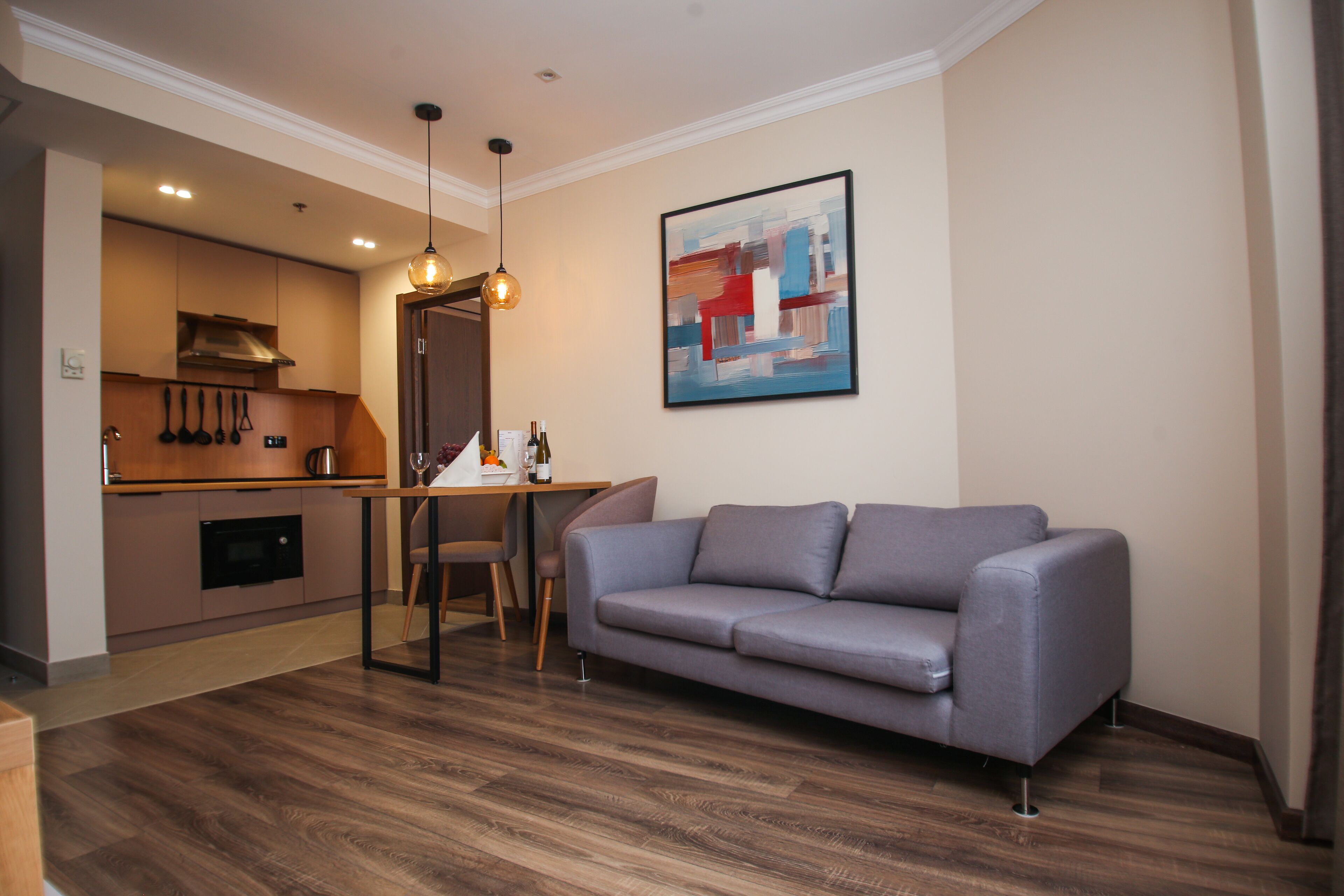 A modern living room with a grey sofa, wooden flooring, a dining table, and a kitchen area with wooden cabinets and a hanging light fixture.