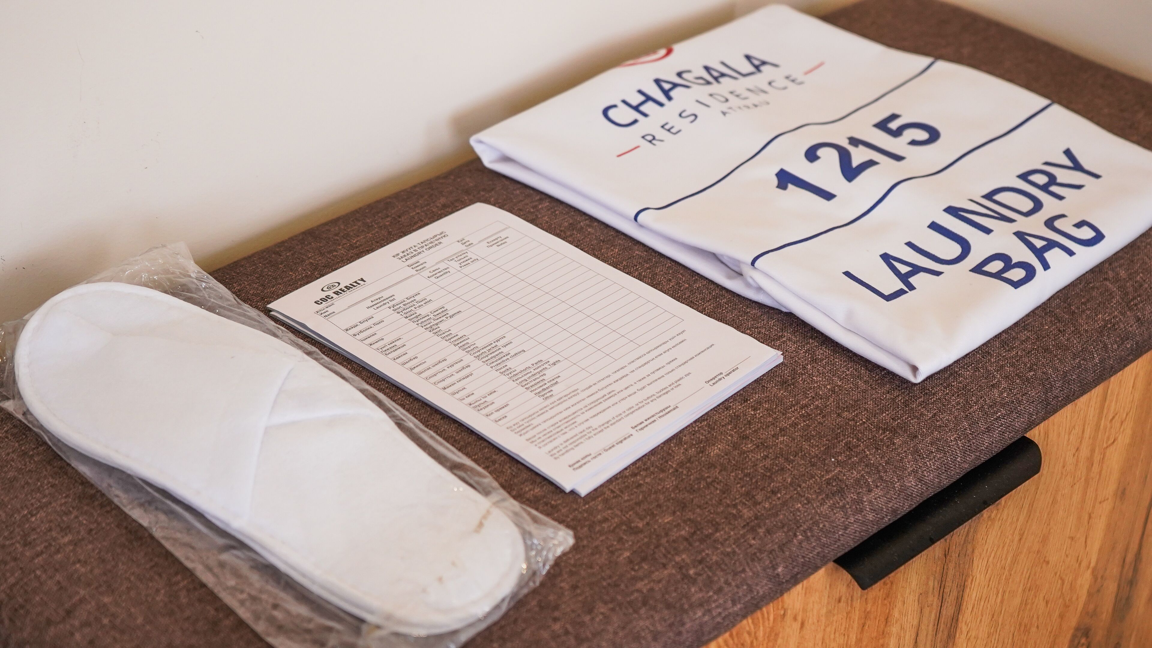Una toalla doblada con la inscripción 'CHASALA RESIDENCE' y '1215 LAUNDRY BAG', un artículo envuelto en plástico y un documento impreso sobre una superficie marrón.