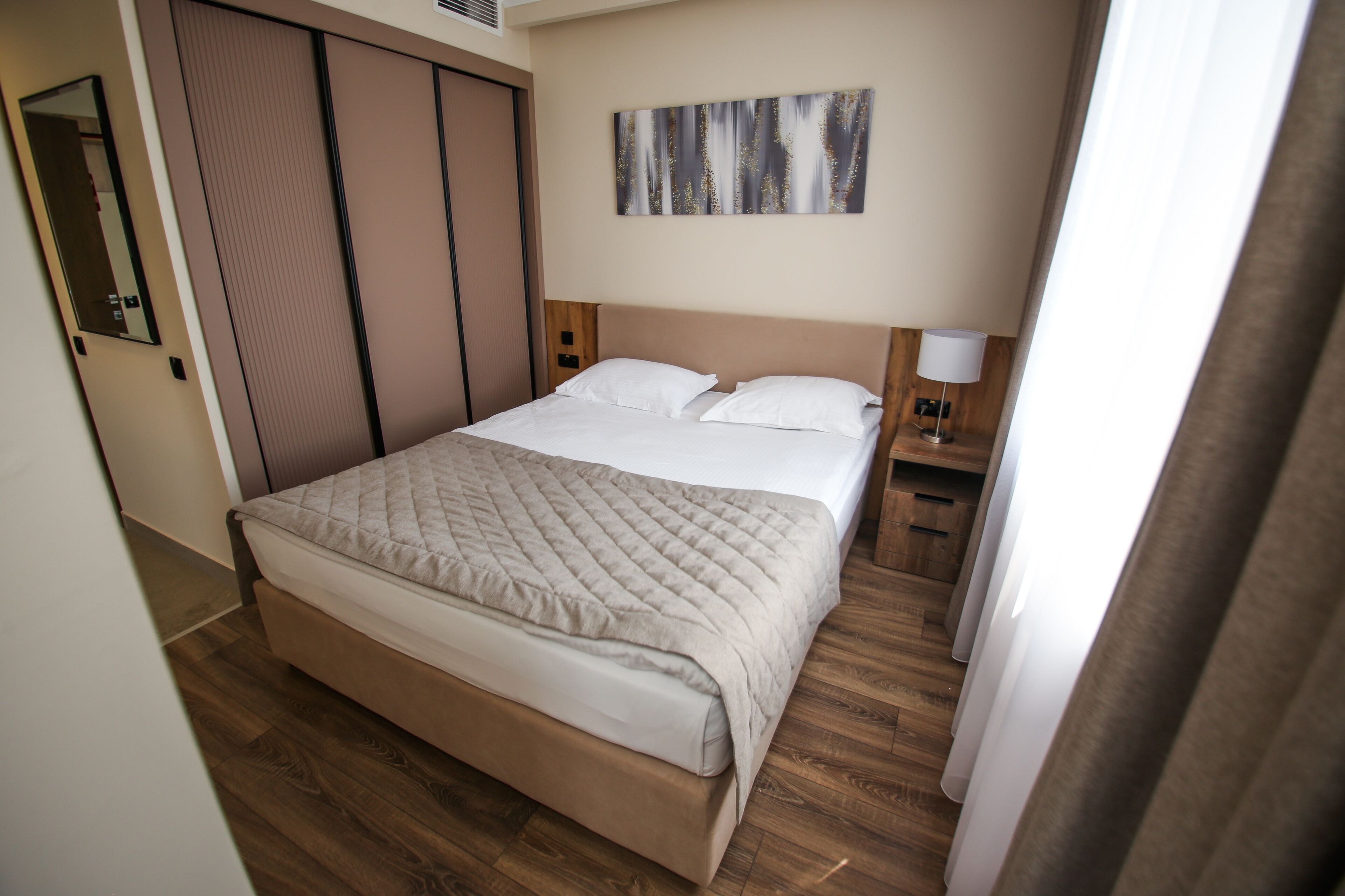 A neatly made bed with a quilted cover, a bedside table with a lamp, a wooden floor, a sliding door closet, and a wall-mounted artwork.