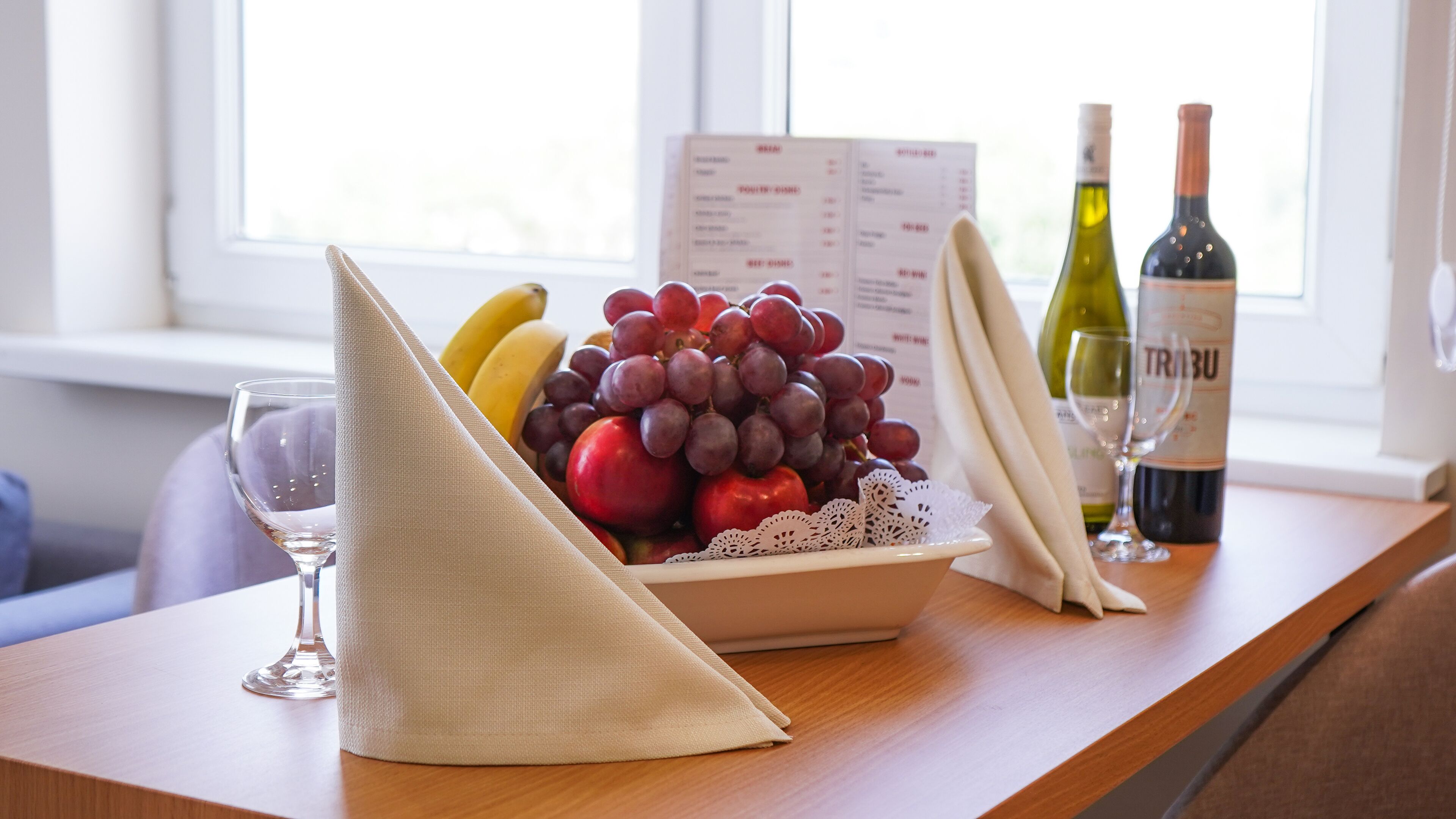 Een tafel met een schaal fruit, een servet, een menukaart en twee flessen wijn.