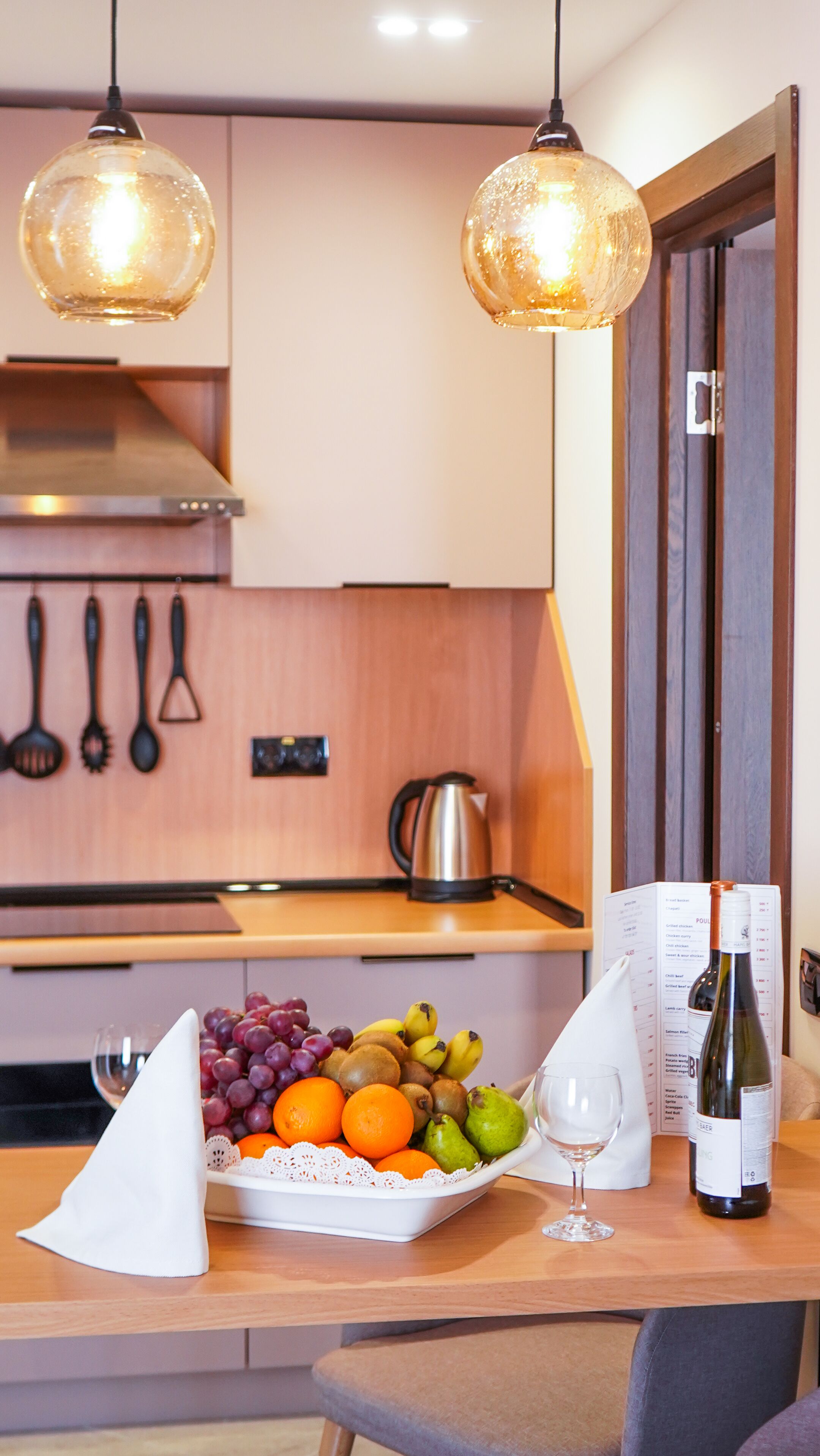 Una cucina moderna con due lampade a sospensione, una ciotola di frutta, una bottiglia di vino e un menu sul tavolo.