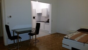 A compact room with a glass-top desk, two chairs, a bed with patterned bedding, and a kitchenette in the background. - Goldfisch Apartment Central Park (Vienna)
