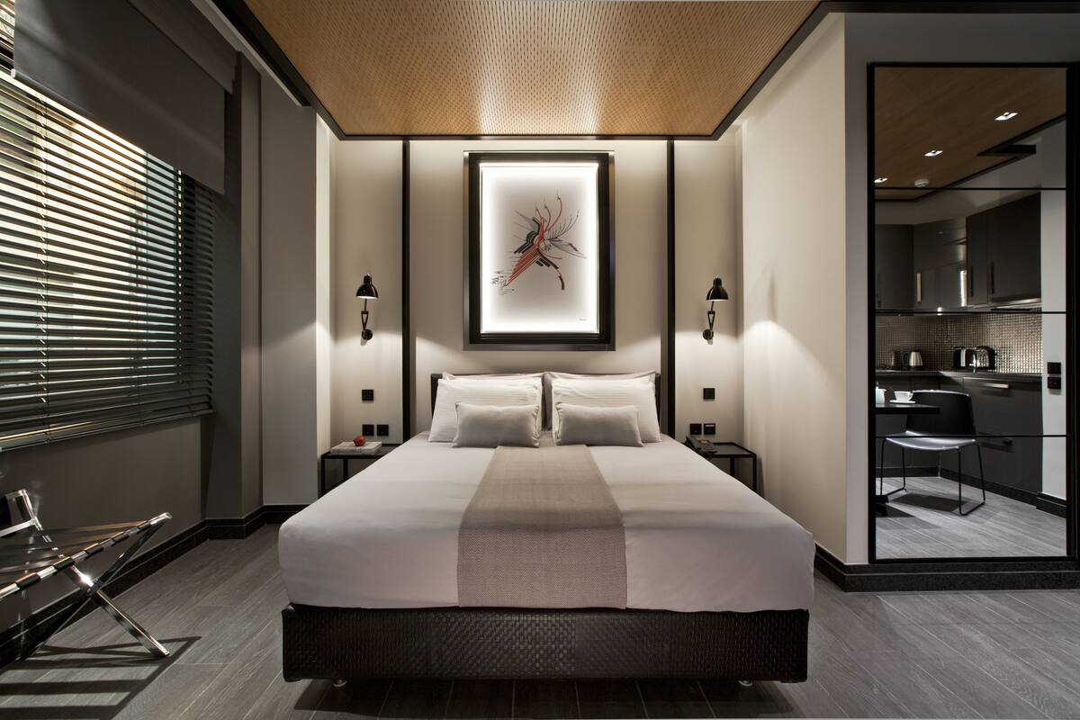 Quarto de hotel moderno com uma cama grande, mesas de cabeceira, uma obra de arte emoldurada e vista para a cozinha.