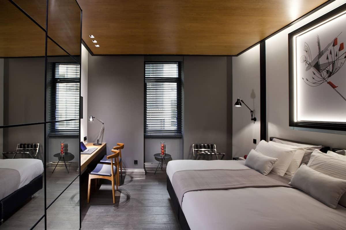 Quarto de hotel moderno com duas camas, uma escrivaninha, uma cadeira, um abajur e um quadro grande em estilo abstrato na parede.