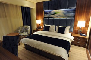 Una camera d'albergo con un letto grande, due comodini con lampade, una sedia e un quadro di grandi dimensioni appeso alla parete.