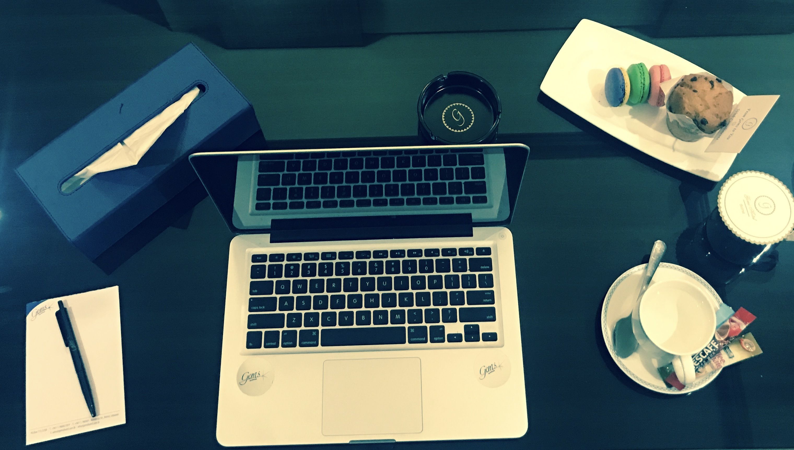 Un ordinateur portable est posé sur une table, à côté d’une tasse, d’une assiette de biscuits, d’un stylo et d’un carnet.