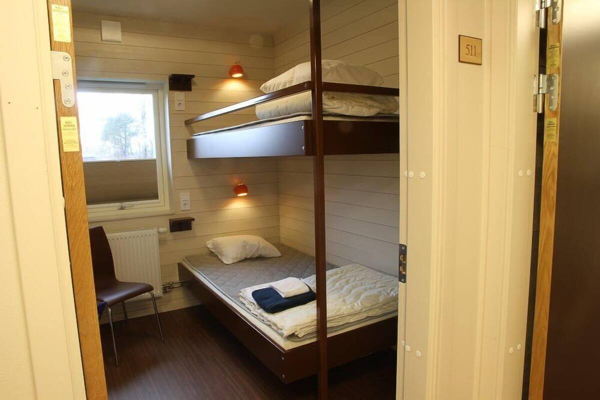 A compact room with bunk beds, a small desk, and a chair. There is a window with blinds and a radiator below it.