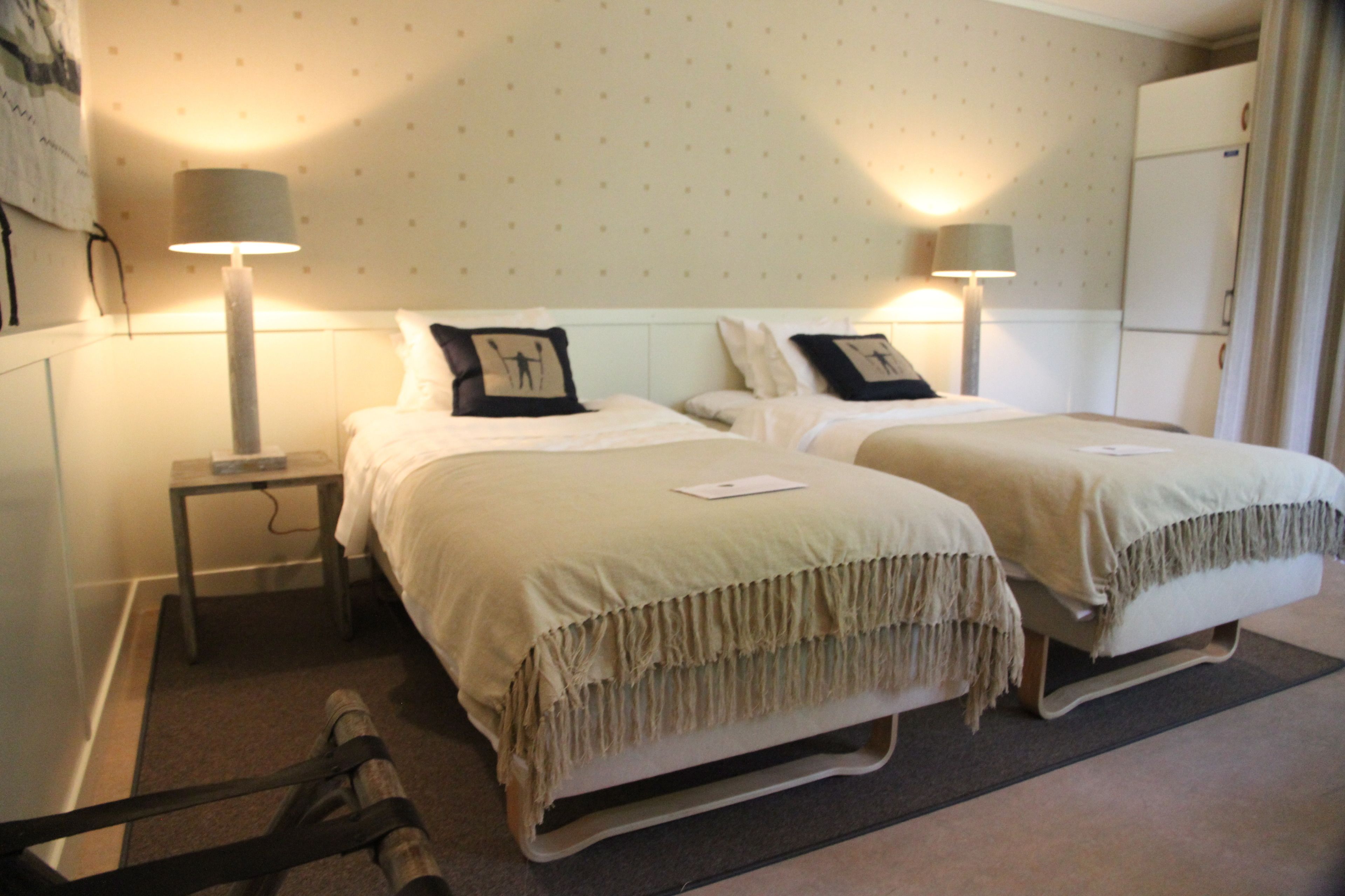 A hotel room with two beds, each with a bedside table and lamp, a small table with a book, and a refrigerator in the corner.
